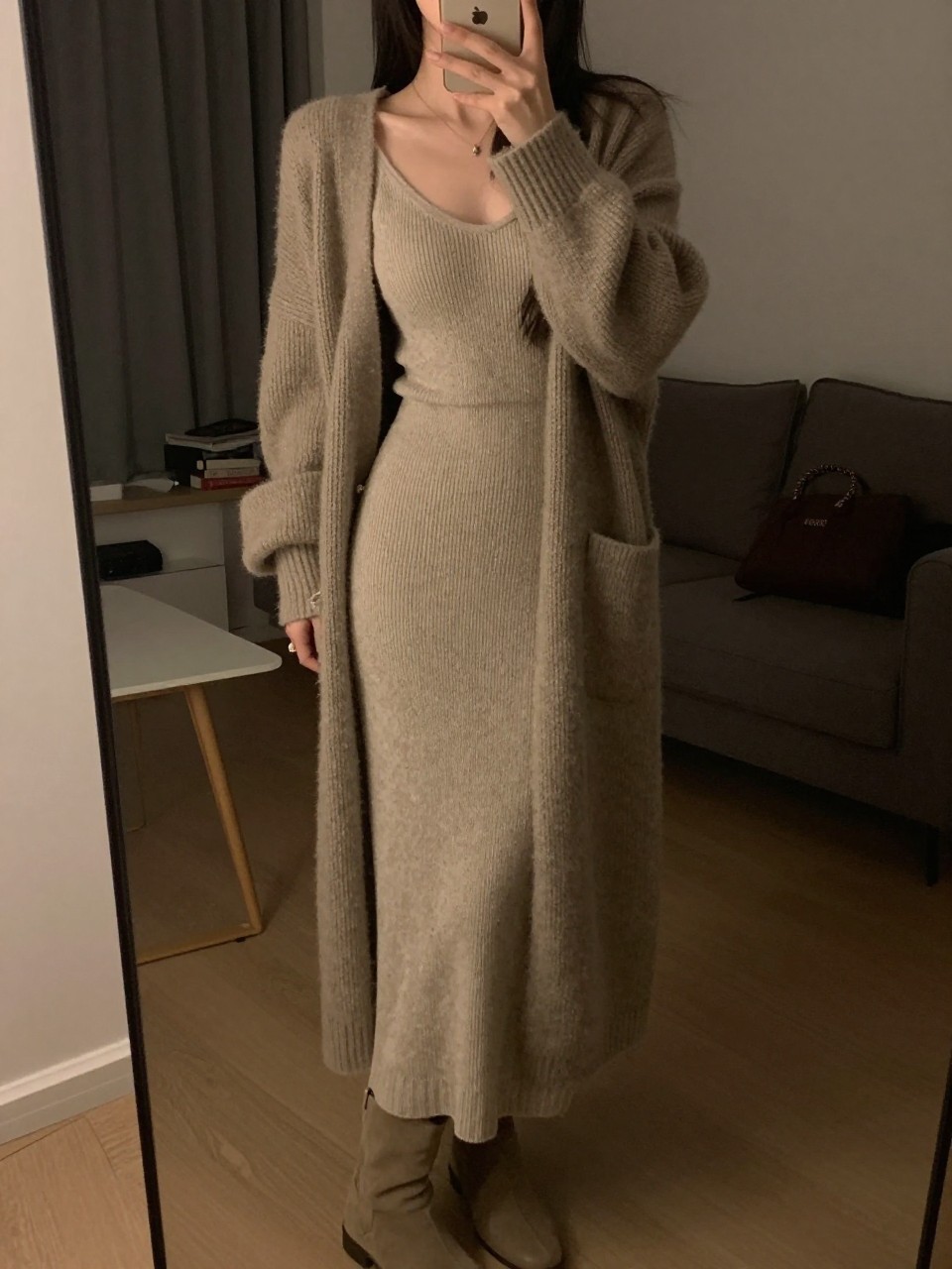 iPhone mirror selfie of a woman in a knit maxi dress with a cashmere cardigan, pulling on boots, cropped at NECK so face doesn't show, in a city apartment, warm moody lighting, iPhone photo quality.