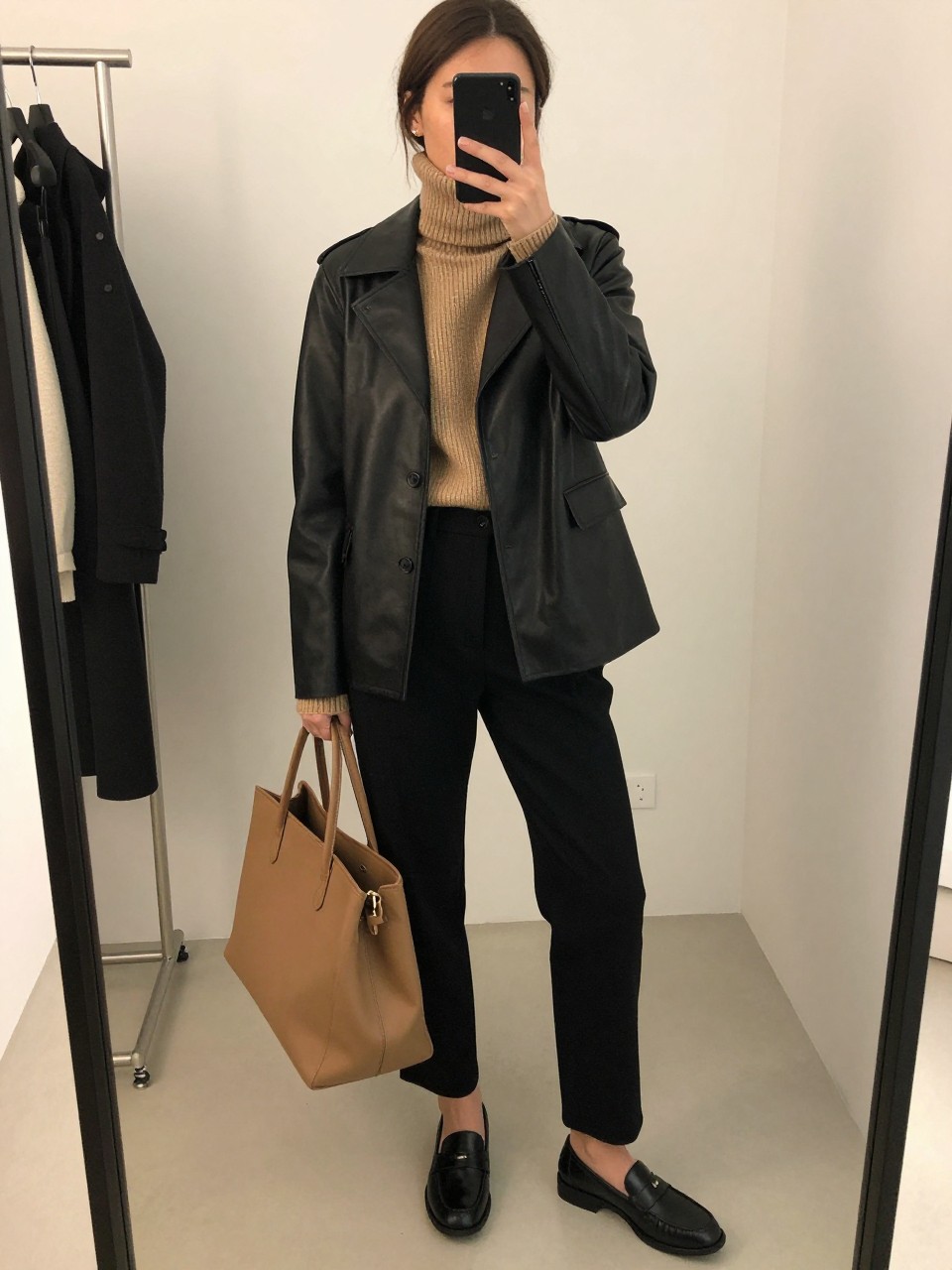 mirror selfie of a brown woman wearing the exact outfit described above, clearly showing a black tailored leather jacket, camel knit turtleneck sweater, black straight-leg trousers, black leather loafers, and a camel structured tote bag, holding a phone in front of a mirror, cropped at NECK so the face does not show, with a relaxed casual selfie pose, in a fashion studio mirror corner, soft indoor lighting, including a clothing rack with garments behind and a tote bag resting on the floor, latest iPhone photo quality.