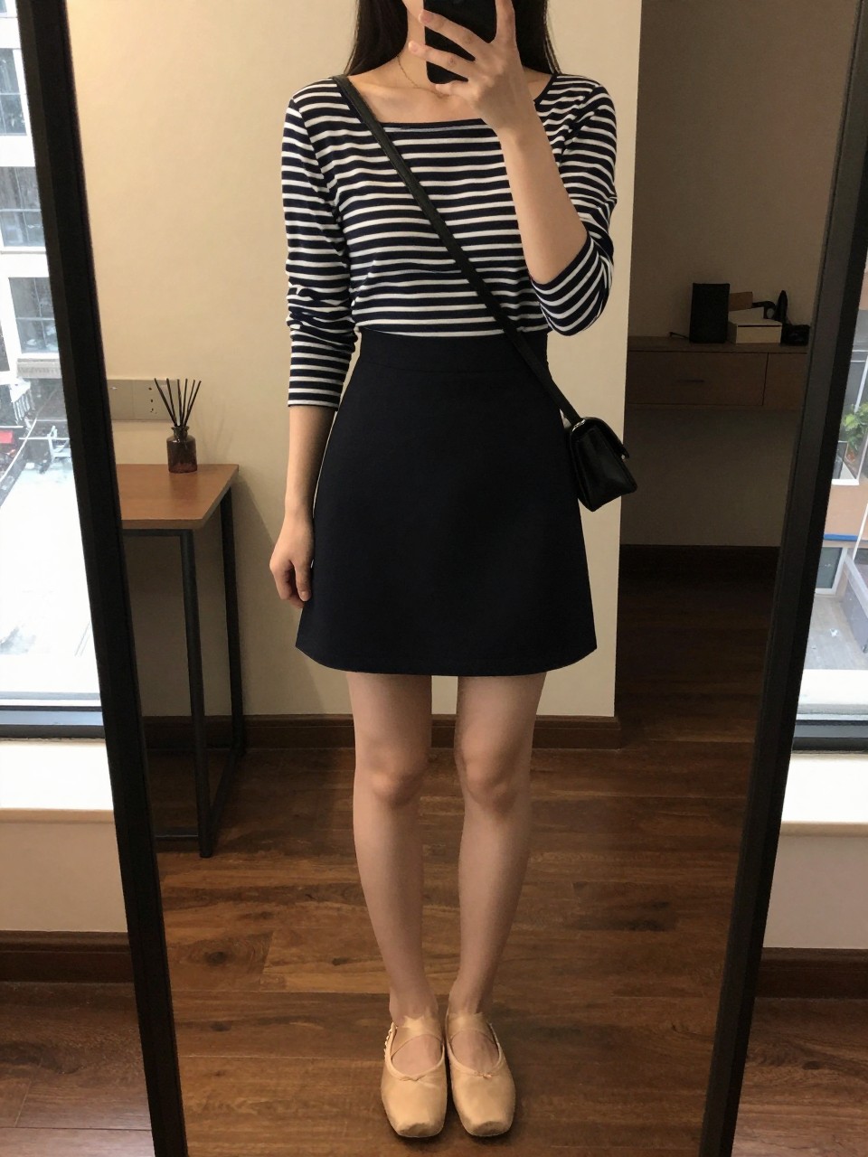 iPhone mirror selfie of a woman in a navy striped boatneck tucked into a black A-line mini skirt with ballet flats, holding a small crossbody bag, cropped at NECK so face doesn't show, in a city apartment, warm moody lighting, iPhone photo quality.
