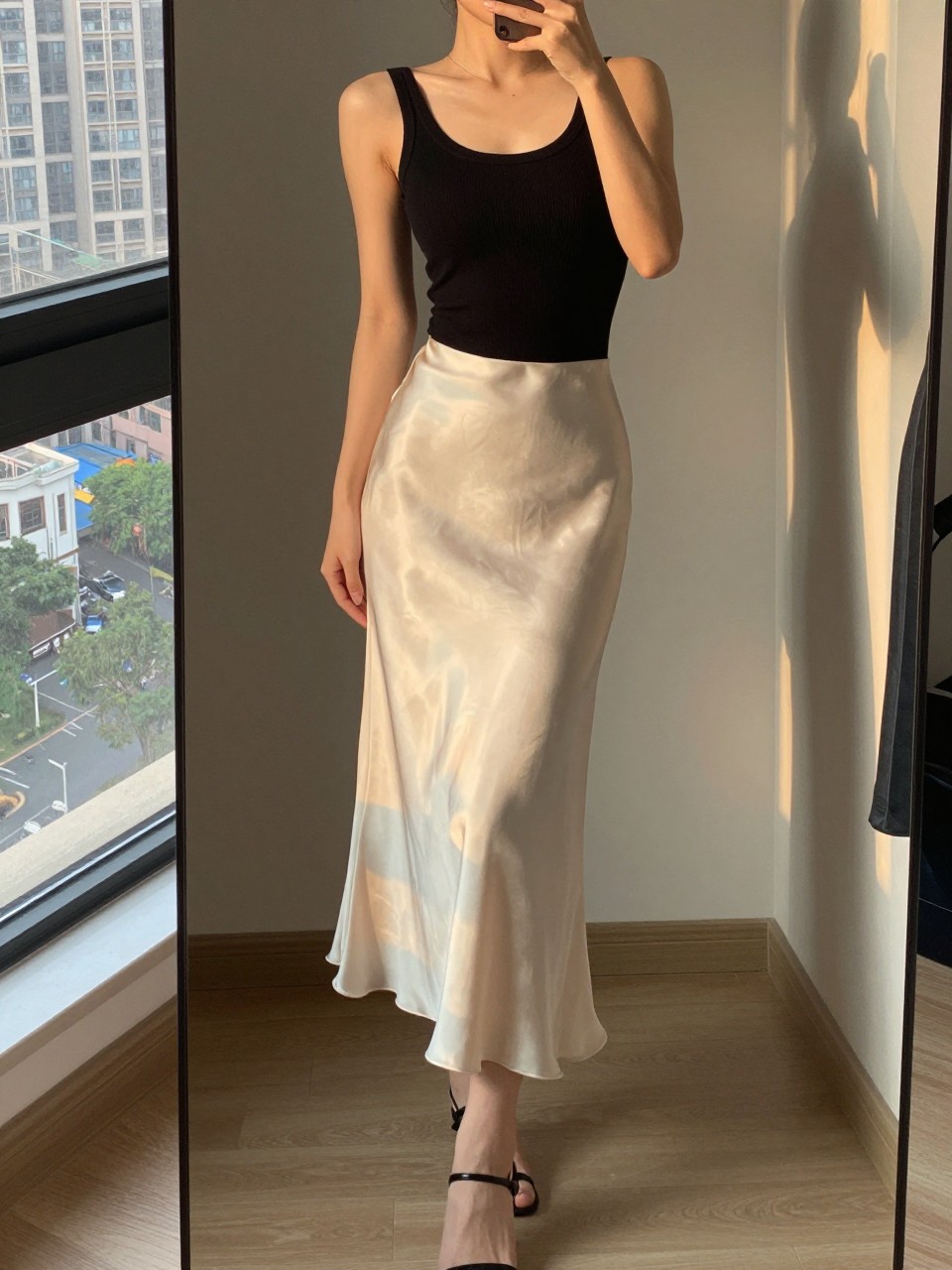 iPhone mirror selfie of a woman wearing an ivory silk slip skirt and a fitted black ribbed tank top with thin-strap heeled sandals, standing sideways to the mirror to show the skirt's drape, cropped at NECK so face doesn't show, in a city apartment, evening golden hour, iPhone photo quality.