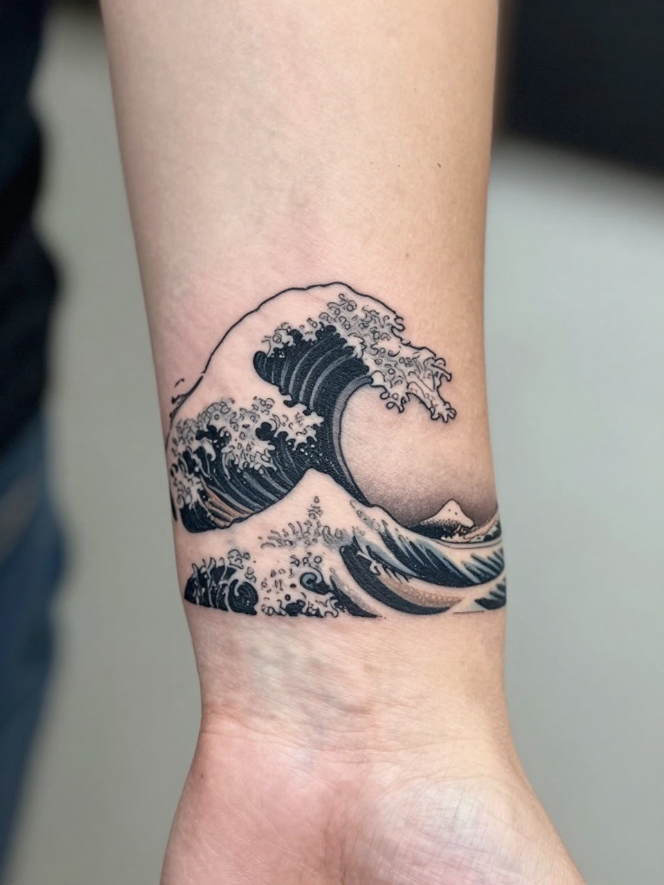 design-focused close-up of a small clean Japanese wave tattoo on the wrist, crisp black ink, fine traditional linework, soft grey shading, minimal composition, realistic skin texture, delicate bracelet nearby, minimal blurred background, modern tattoo inspiration photo (no watermarks on images)