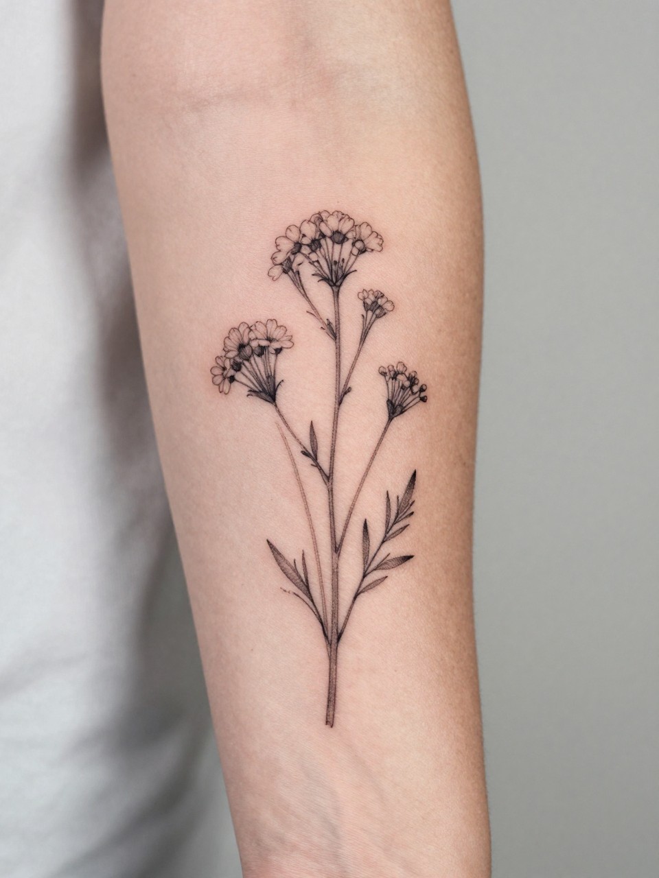 design-focused close-up of a wildflower stem tattoo on women's inner arm, black ink, fine line leaves and tiny blooms, clean earthy boho hippie style, realistic skin texture, delicate ring visible, minimal soft background, feminine modern tattoo inspiration photo (no watermarks on images)