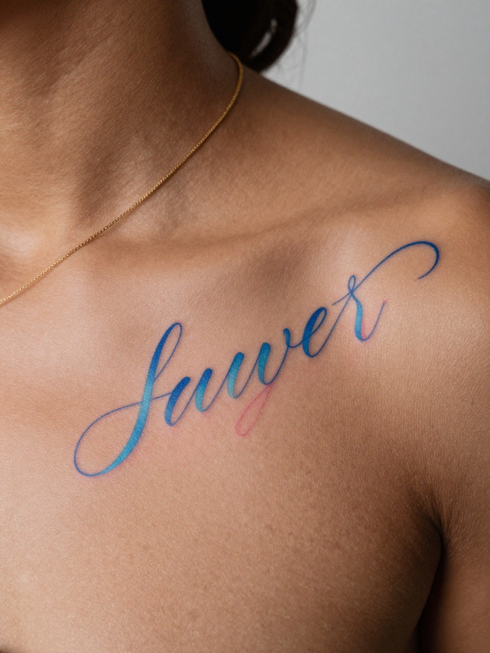 close-up of a blue and pink script tattoo on rich brown skin at the collarbone, elegant cursive lettering, soft pastel blue and pink tones, clean outline, realistic skin texture, gold necklace chain visible, design-focused close-up, minimal blurred background, modern tattoo photography (no watermarks on images)