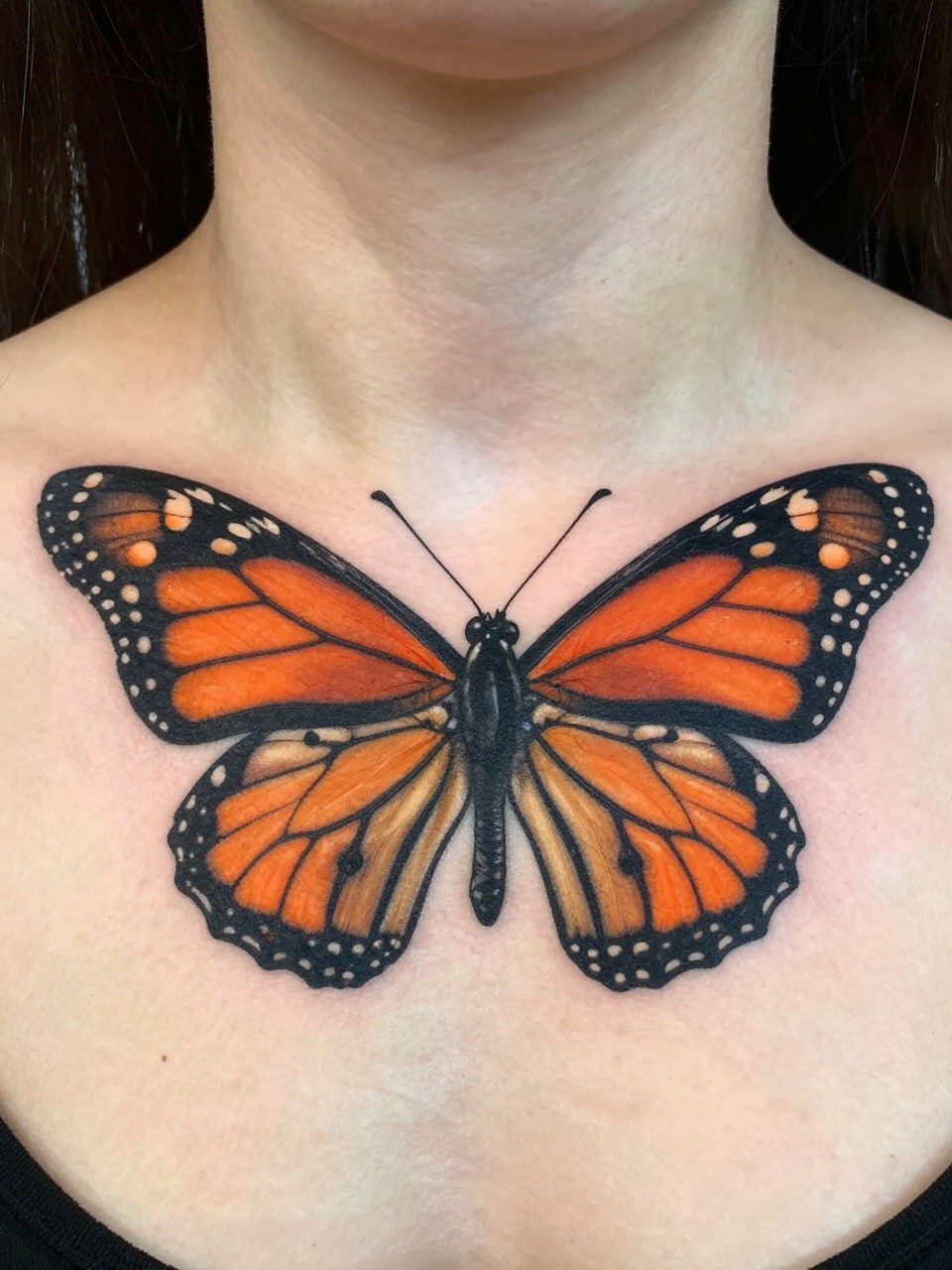 "Close-up of the collarbone and lower neck: a large, richly colored monarch butterfly tattoo centered on the sternum, its upper wings reaching toward the throat. Bold lines, vivid orange, stark against clean skin."