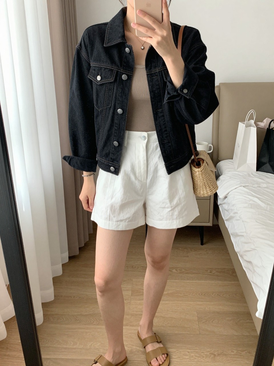 mirror selfie of a woman wearing the exact outfit described above, clearly showing a black slightly boxy denim jean jacket, taupe fitted tank top, white tailored linen shorts, tan slide sandals, and a straw crossbody bag, holding a phone in front of a mirror, cropped at NECK so the face does not show, with a relaxed casual selfie pose, in a feminine-themed bedroom, natural window light, including a coffee mug on a bedside table and shopping bags beside a chair, latest iPhone photo quality.