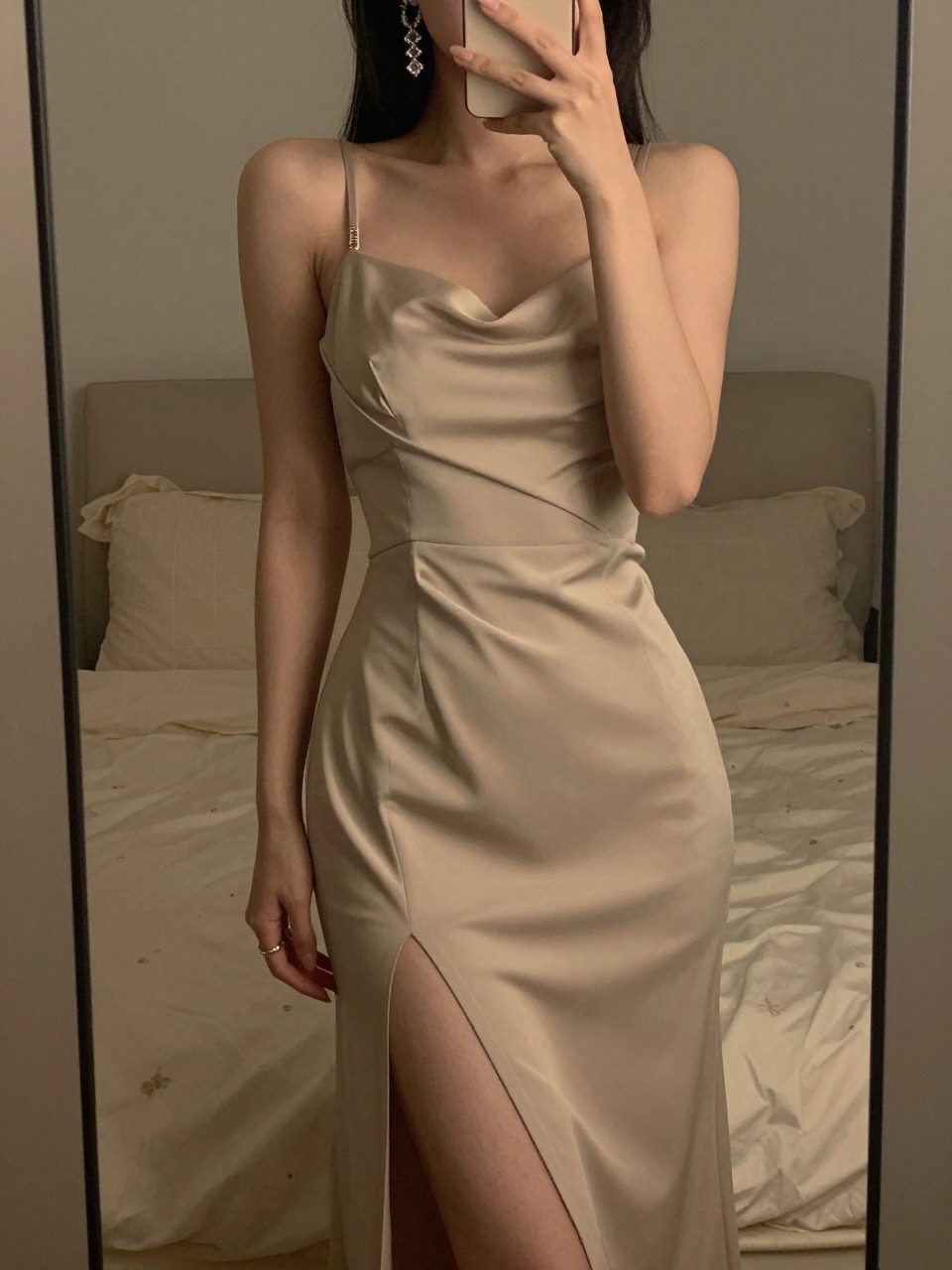 iPhone mirror selfie of a woman in a high-slit gown and statement earrings, posing gracefully, cropped at the neck so face doesn't show, in a feminine-themed bedroom, warm moody lighting, iPhone photo quality.
