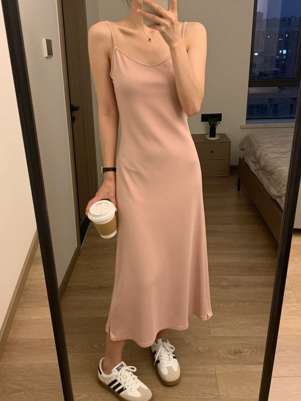 iPhone mirror selfie of a woman in a baby pink slip dress and white sneakers, holding a coffee cup, cropped at the neck so face doesn't show, in a city apartment, warm moody lighting, iPhone photo quality.