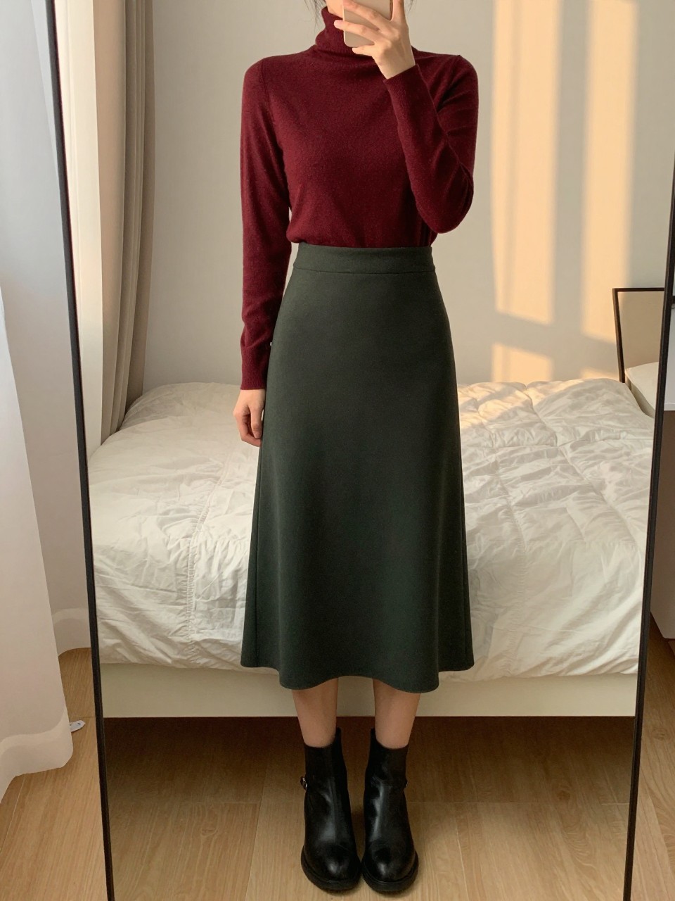 iPhone mirror selfie of a woman in a burgundy turtleneck tucked into a forest green midi skirt with black ankle boots, standing with weight shifted, cropped at NECK so face doesn't show, in a feminine-themed bedroom, evening golden hour, iPhone photo quality.
