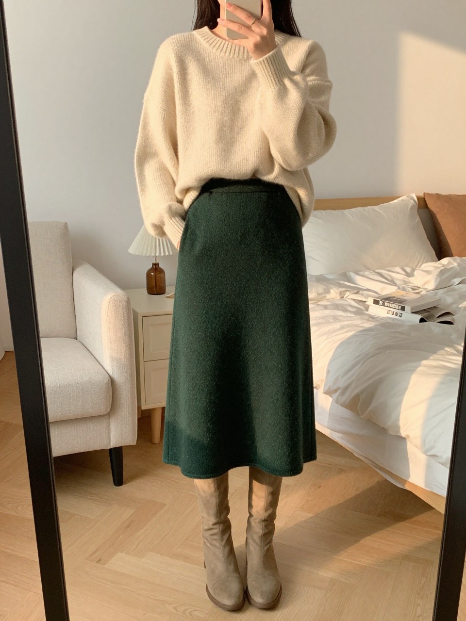 iPhone mirror selfie of a woman in an oversized cream sweater and forest green wool midi skirt with tan knee-high boots, hands in sweater pockets, cropped at NECK so face doesn't show, in a feminine-themed bedroom, evening golden hour, iPhone photo quality.