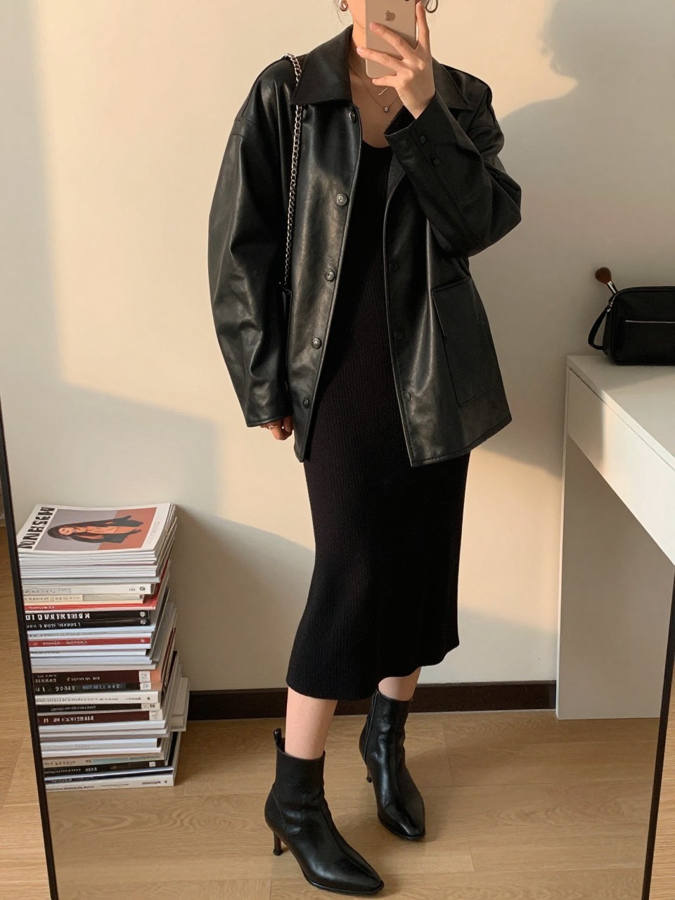 mirror selfie of a woman wearing the exact outfit described above, clearly showing an oversized black leather jacket with dropped shoulders, black knit midi dress in soft stretch fabric, black ankle boots with a slim heel, silver chain-strap bag, and small silver hoop earrings, holding a phone in front of a mirror, cropped at NECK so the face does not show, with a relaxed casual selfie pose, in a minimal dressing room, evening golden hour, including stack of fashion magazines on a side table and a makeup brush or cosmetic bag on the counter, latest iPhone photo quality.