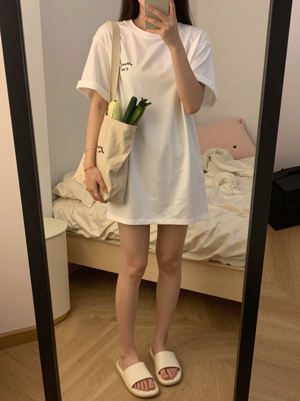 iPhone mirror selfie of a woman in a white t-shirt dress and slide sandals, holding a bag of produce, cropped at the neck so face doesn't show, in a feminine-themed bedroom, warm moody lighting, iPhone photo quality.