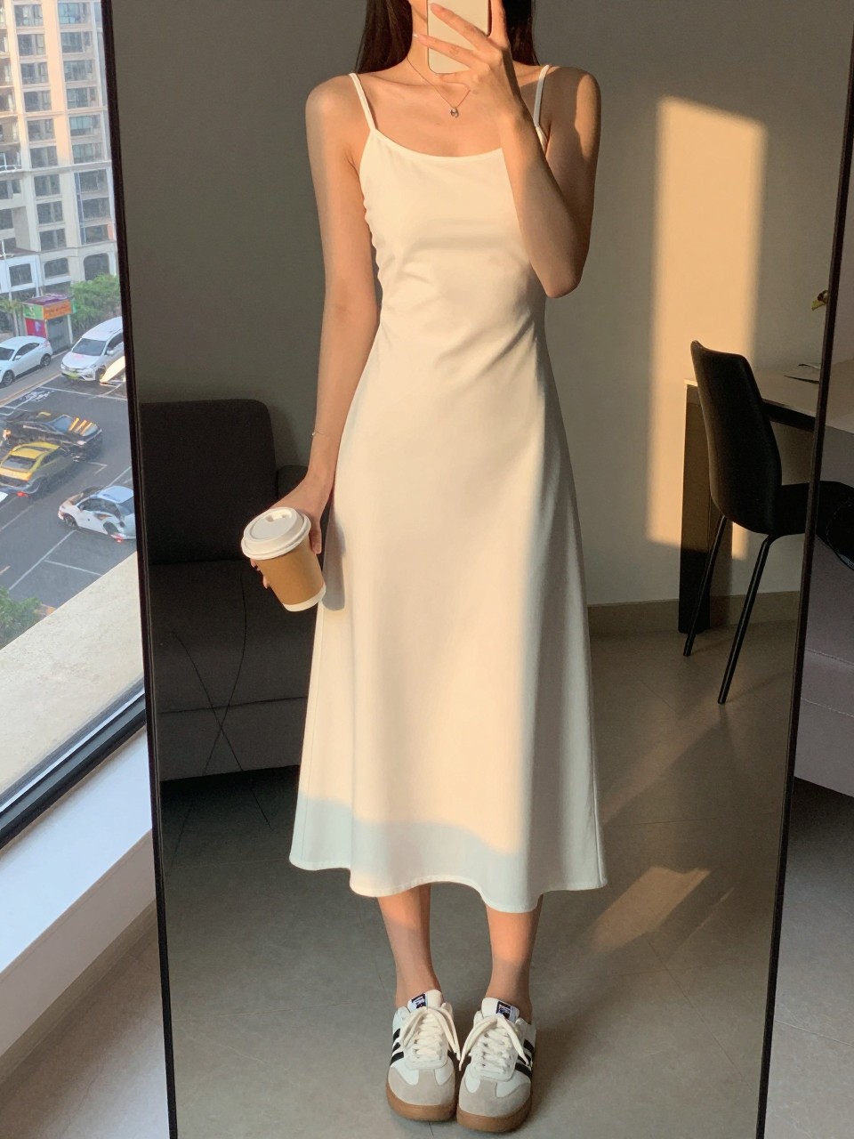 iPhone mirror selfie of a woman in a white shift dress and sneakers, holding a coffee cup, cropped at the neck so face doesn't show, in a city apartment, evening golden hour lighting, iPhone photo quality.