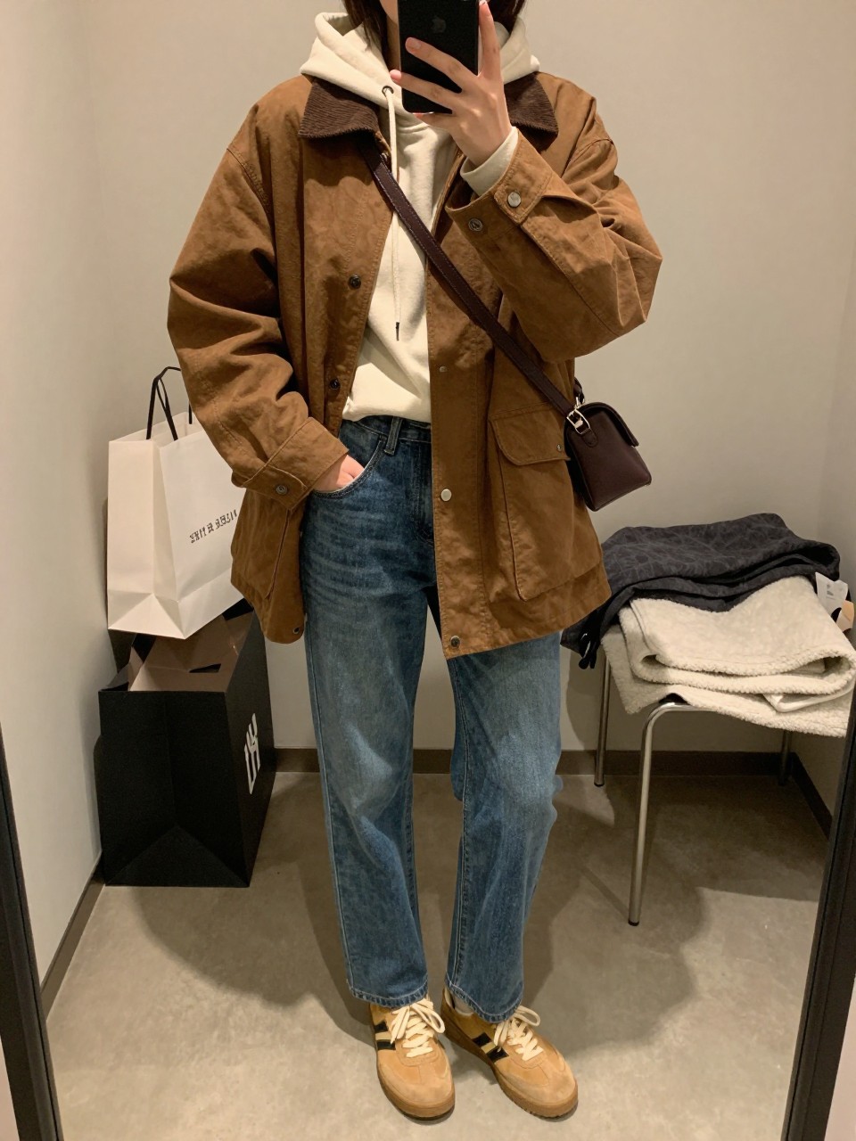 mirror selfie of a woman wearing the exact outfit described above, clearly showing a brown canvas barn jacket with a relaxed fit and corduroy collar, cream hoodie in soft sweatshirt fabric, blue straight-leg jeans, tan suede sneakers, and a dark brown belt bag, holding a phone in front of a mirror, cropped at NECK so the face does not show, with a relaxed casual selfie pose, in a walk-in closet mirror, warm moody lighting, including shopping bags beside a chair and folded blanket on a chair, latest iPhone photo quality.