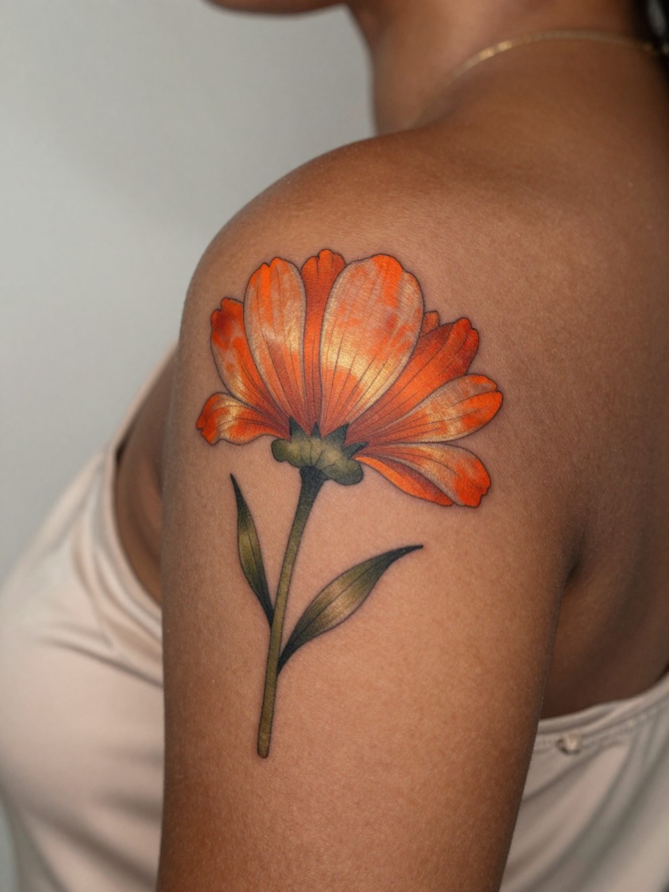 close-up of a sunset flower tattoo on deep brown skin at the shoulder, orange coral and golden petals, soft green stem detail, clean outline, smooth shading, realistic skin texture, silk blouse edge visible, design-focused close-up, neutral blurred background, modern tattoo photography (no watermarks on images)