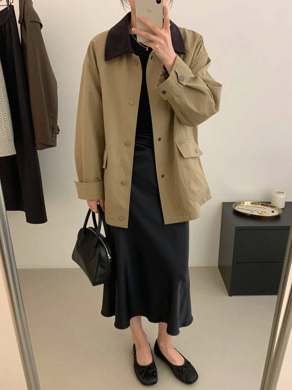 mirror selfie of a woman wearing the exact outfit described above, clearly showing a khaki barn jacket with a dark corduroy collar, black fitted long-sleeve top, black satin midi skirt, black ballet flats, and a small structured black handbag, holding a phone in front of a mirror, cropped at NECK so the face does not show, with a relaxed casual selfie pose, in a fashion studio mirror corner, soft indoor lighting, including a clothing rack with garments behind and a jewelry tray on the vanity, latest iPhone photo quality.