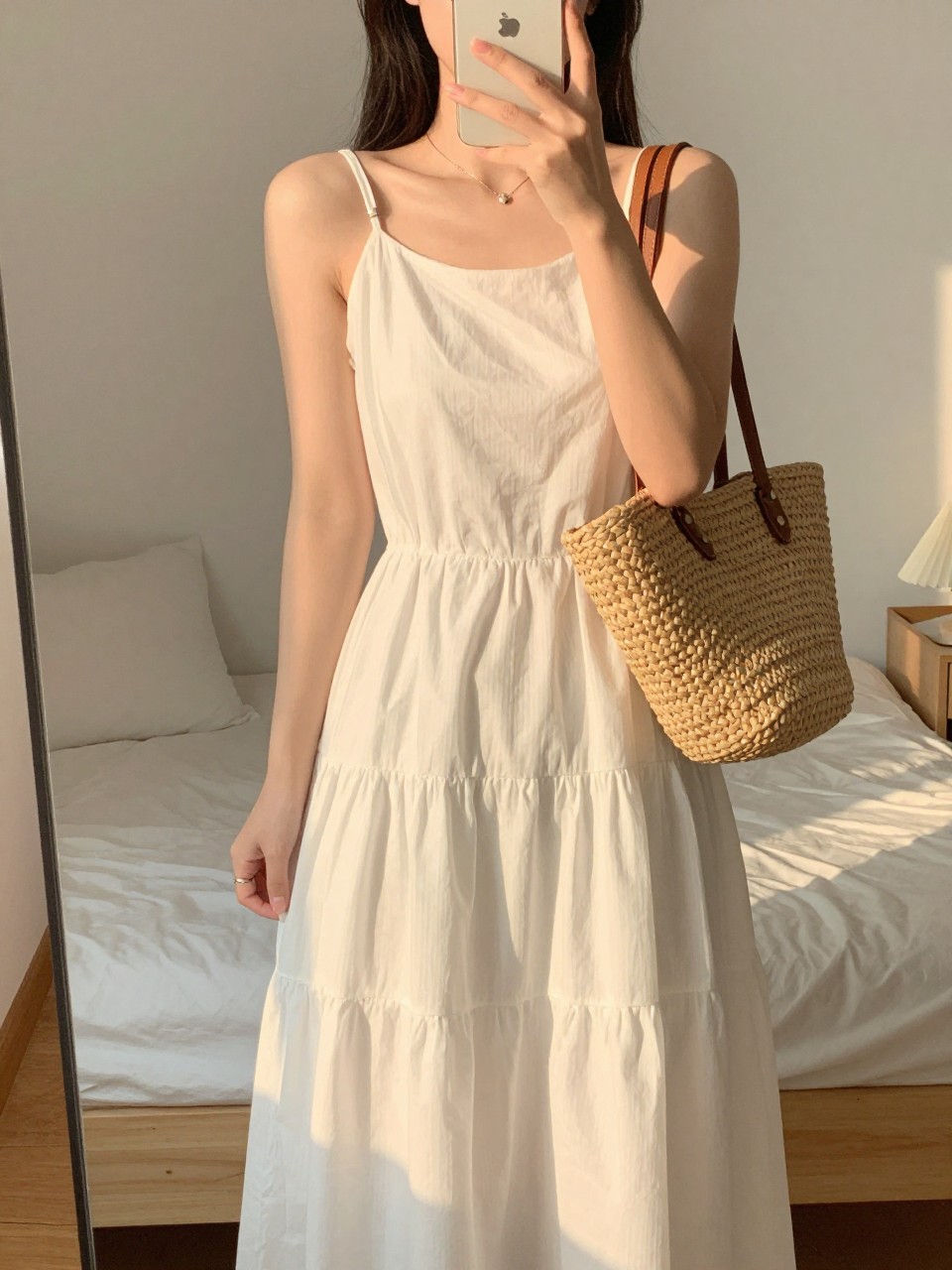 iPhone mirror selfie of a woman in a white tiered dress holding a straw bag, laughing in the sunlight, cropped at the neck so face doesn't show, in a feminine-themed bedroom, evening golden hour lighting, iPhone photo quality.