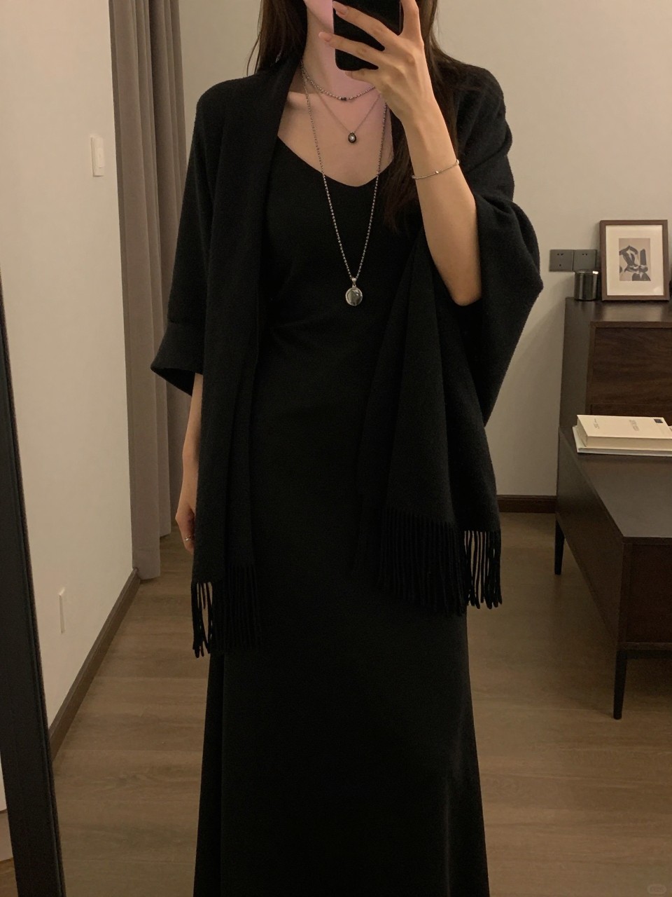 iPhone mirror selfie of a woman in a black maxi with a fringed shawl and layered necklaces, posing softly, cropped at NECK so face doesn't show, in a city apartment, warm moody lighting, iPhone photo quality.