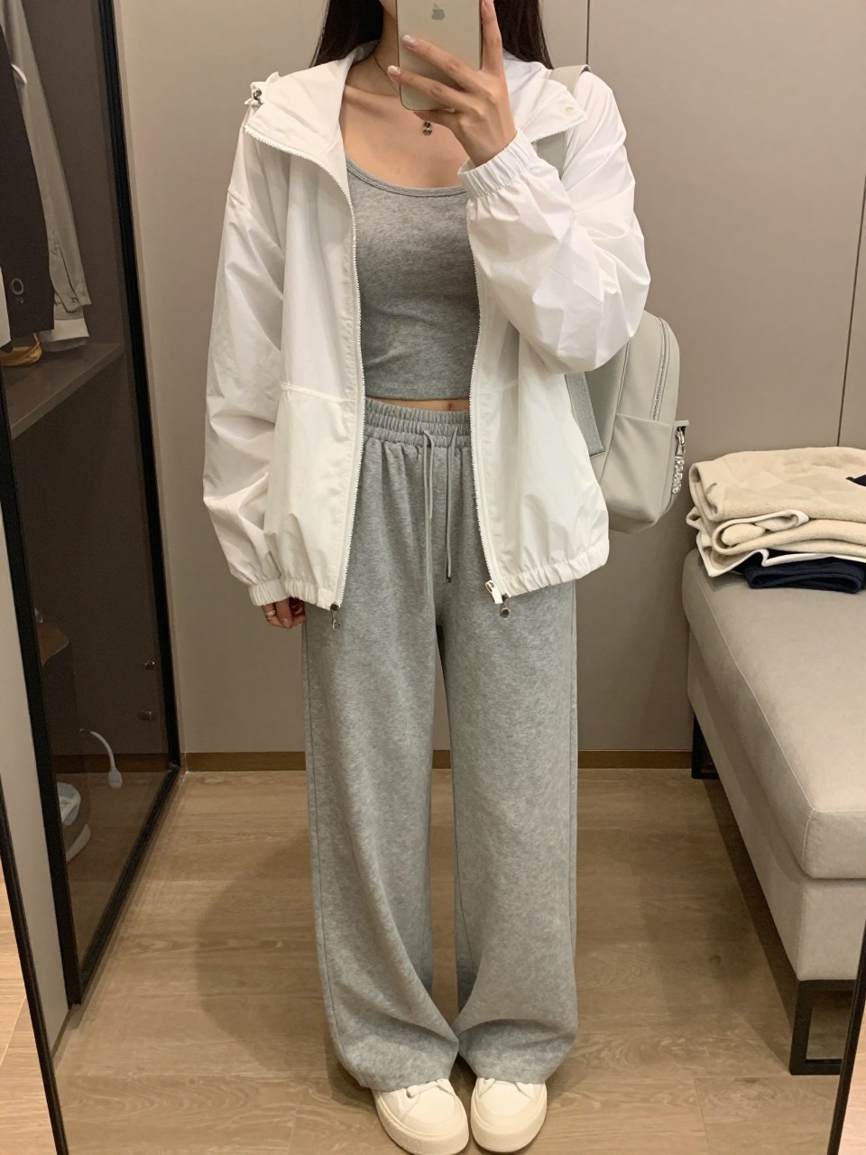 mirror selfie of a woman wearing a white lightweight windbreaker with a clean zip front, a heather gray fitted crop top, soft gray wide-leg sweatpants, white platform sneakers, and a pale gray mini backpack, holding a phone in front of a mirror, cropped at NECK so the face does not show, with a relaxed casual selfie pose, in a walk-in closet mirror, soft indoor lighting, including an open closet with clothes visible and a folded blanket on a chair, latest iPhone photo quality. (no watermarks on images)