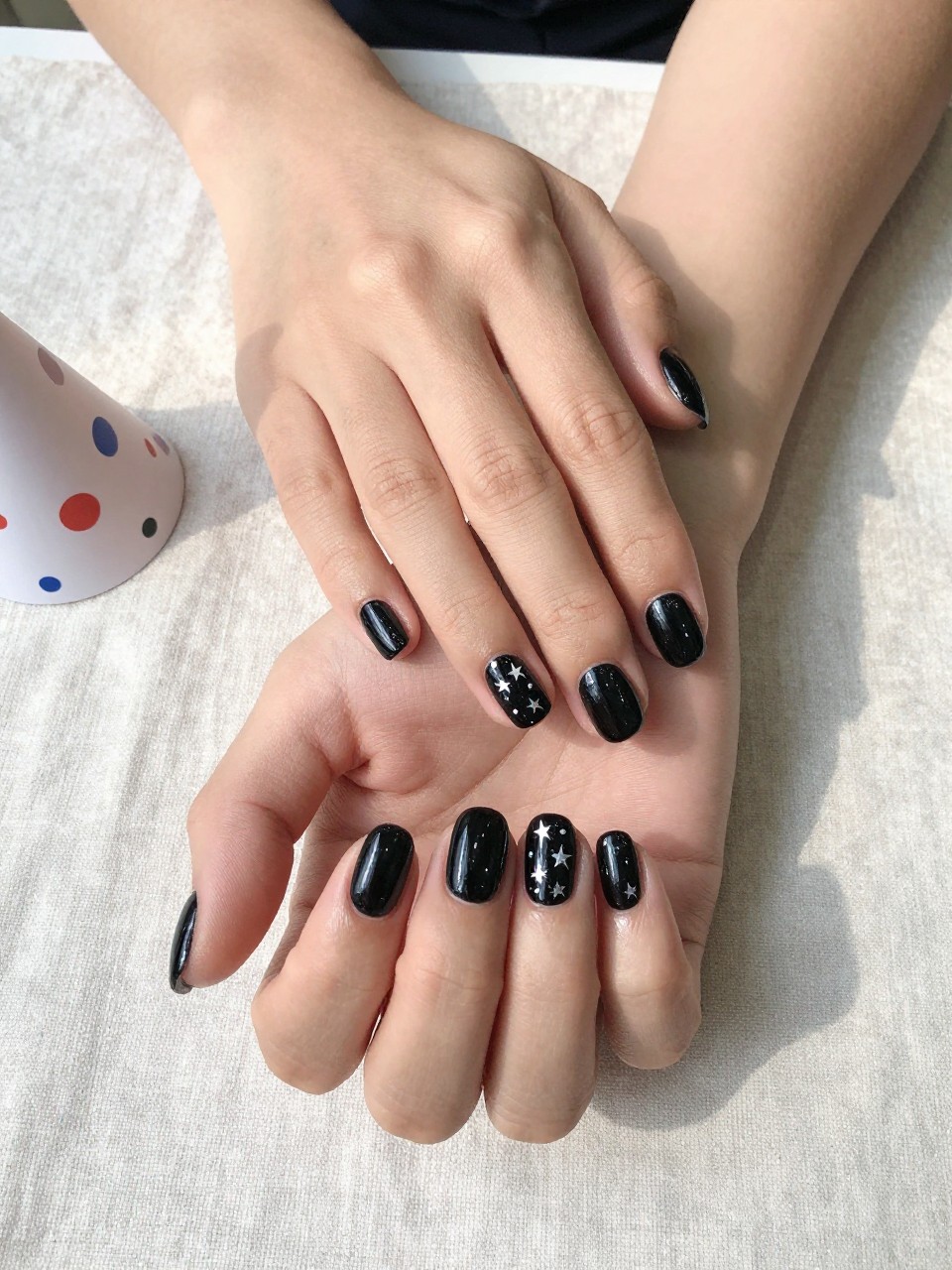 90-degree overhead top-down view of two feminine hands with oval medium nails.
The manicure features a glossy black base with tiny silver star accents scattered across two accent nails.
Include glossy gel finish.

Hands are posed one hand resting lightly over the other.

Scene set on a soft linen cloth with a small party hat nearby.

Bright natural daylight with soft shadows, hyper-realistic lifestyle photography, sharp focus on nails, natural skin texture, realistic hands, no extra fingers.
