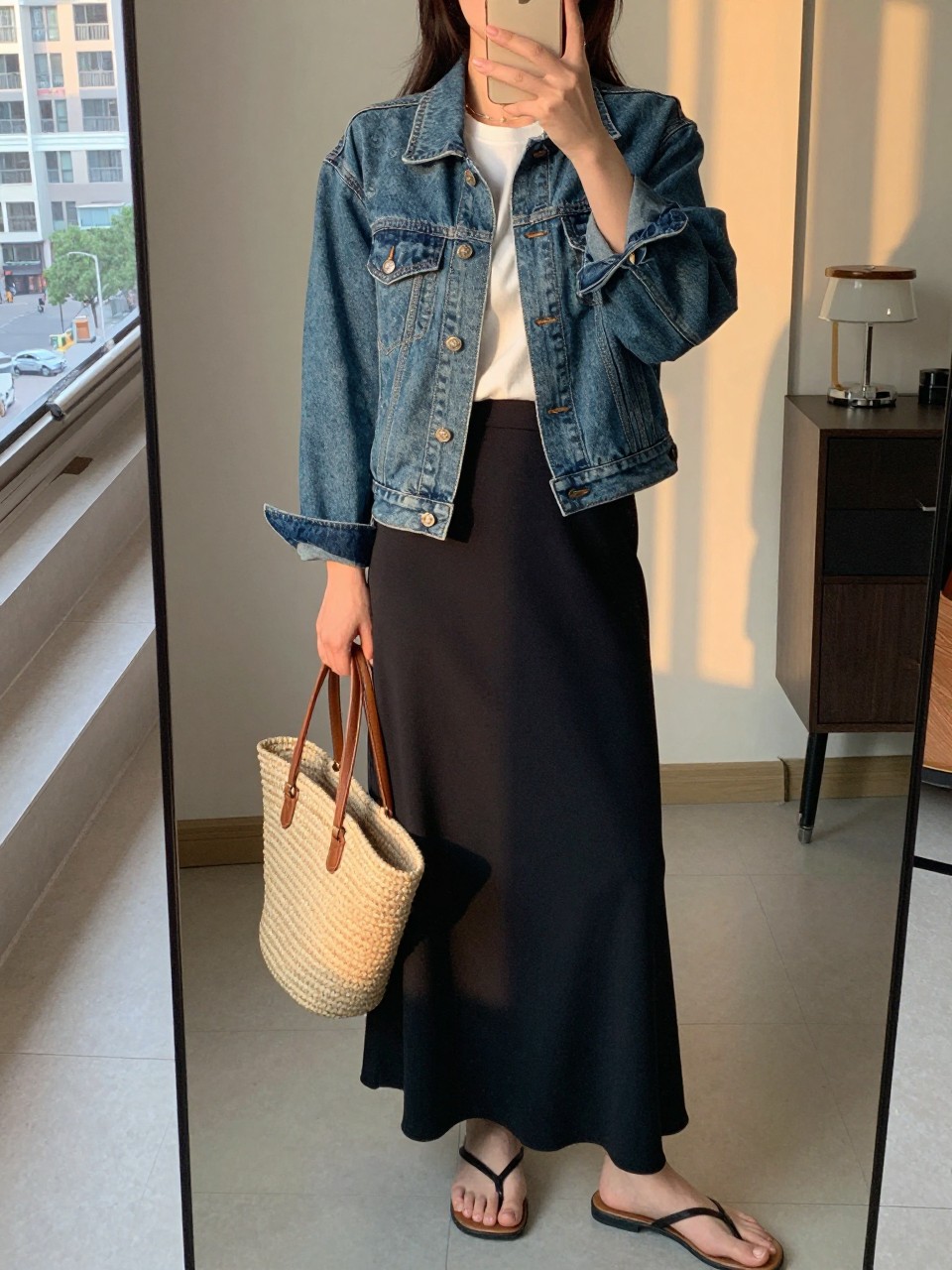 iPhone mirror selfie of a woman in a denim jacket over a white tee and black maxi skirt with flat sandals, holding a straw tote, cropped at NECK so face doesn't show, in a city apartment, evening golden hour, iPhone photo quality.
