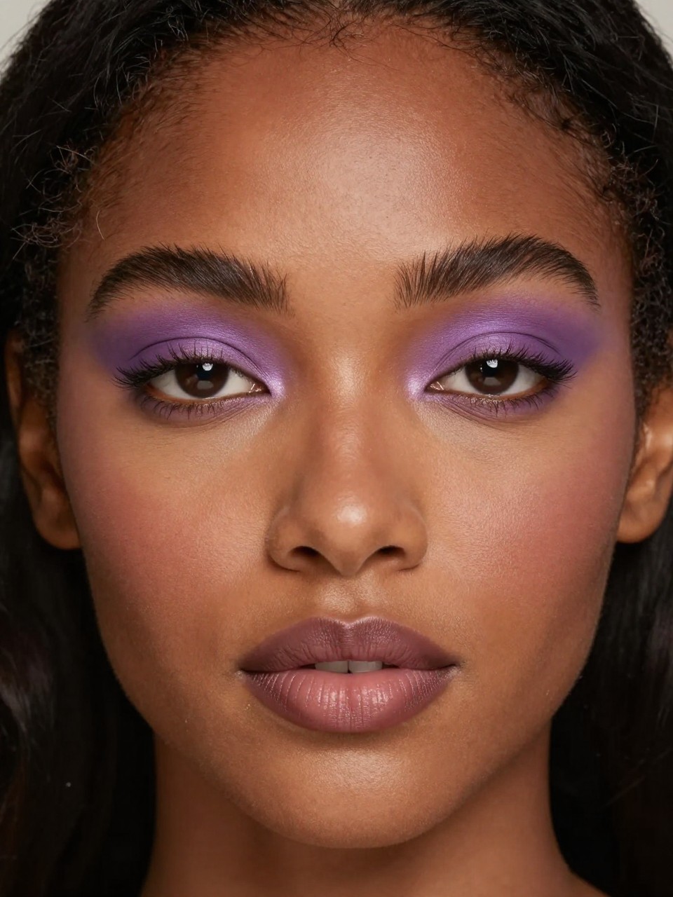 Super realistic close-up beauty portrait of a black woman wearing dusty purple matte eyeshadow blended smoothly over the lids with muted brown outer corners, soft matte skin, lightly bronzed cheeks, fluffy brushed-up brows, muted mauve lips, and defined natural lashes, shot from a front-facing portrait, with diffused studio lighting that clearly reveals the matte texture and skin finish, realistic skin texture, softly styled hair framing the face, minimal neutral background, feminine modern beauty photography, latest iPhone photo quality (no watermarks on images).