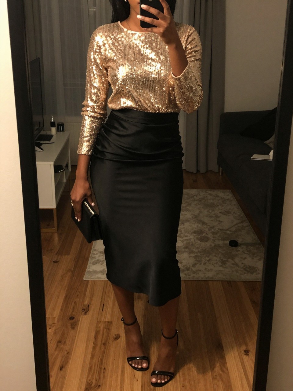 iPhone mirror selfie of a black woman in a gold sequin top tucked into a black satin midi skirt with strappy heels, holding a mini clutch, cropped at NECK so face doesn't show, in a city apartment, warm moody lighting, iPhone photo quality.