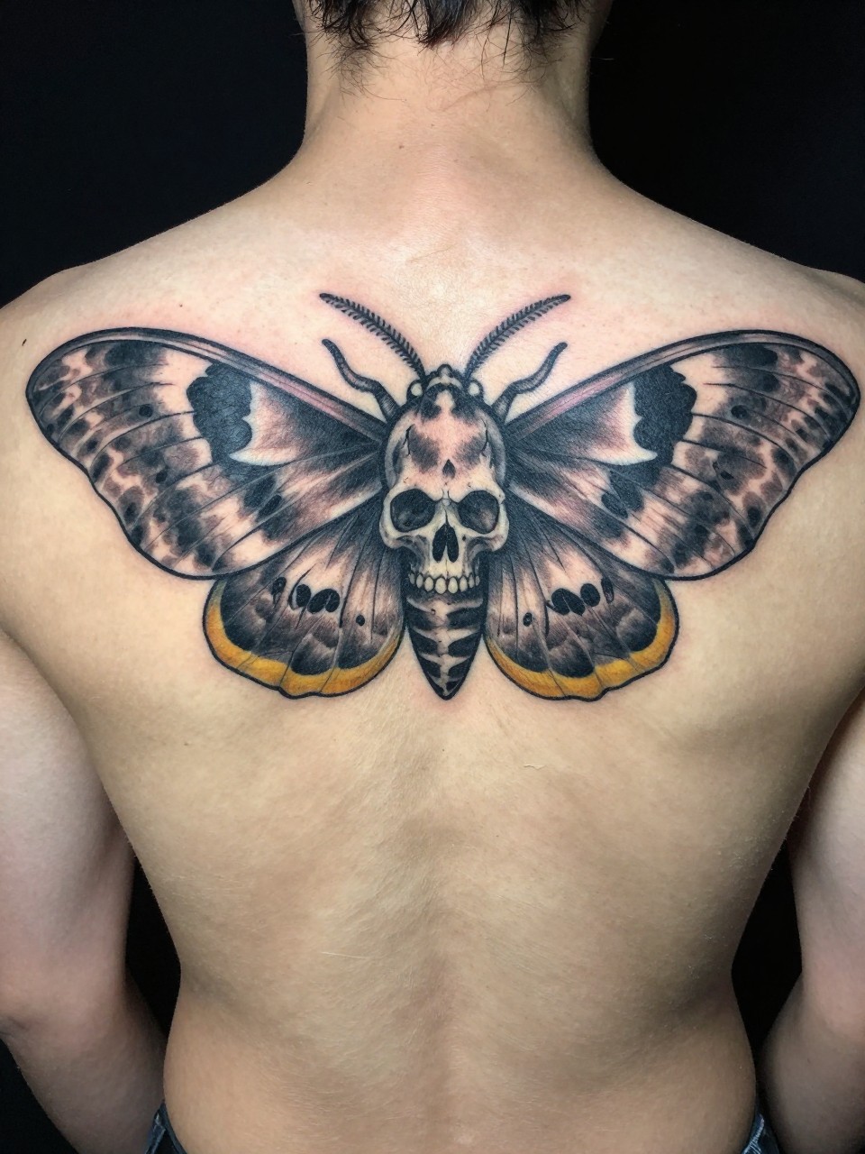 Full back of neck/upper back: a large death's-head moth tattoo where the skull pattern is exaggerated, and the lower wings fade into a reaper's cloak folds."