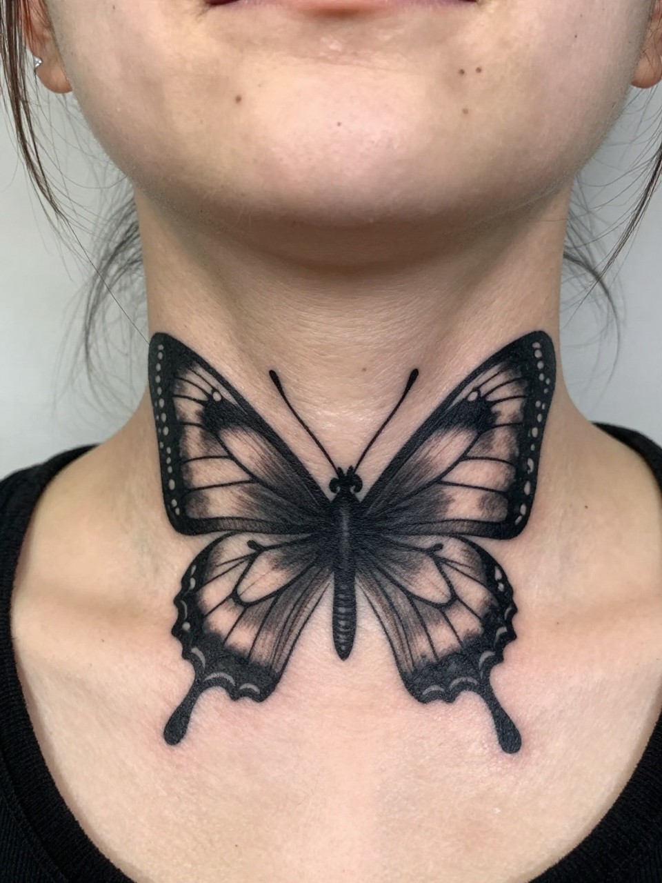  "Low-angle close-up from the fron of the neckt: a bold, graphic butterfly tattoo centered under the chin. The perspective is bold and unmissable, with strong blackwork."
