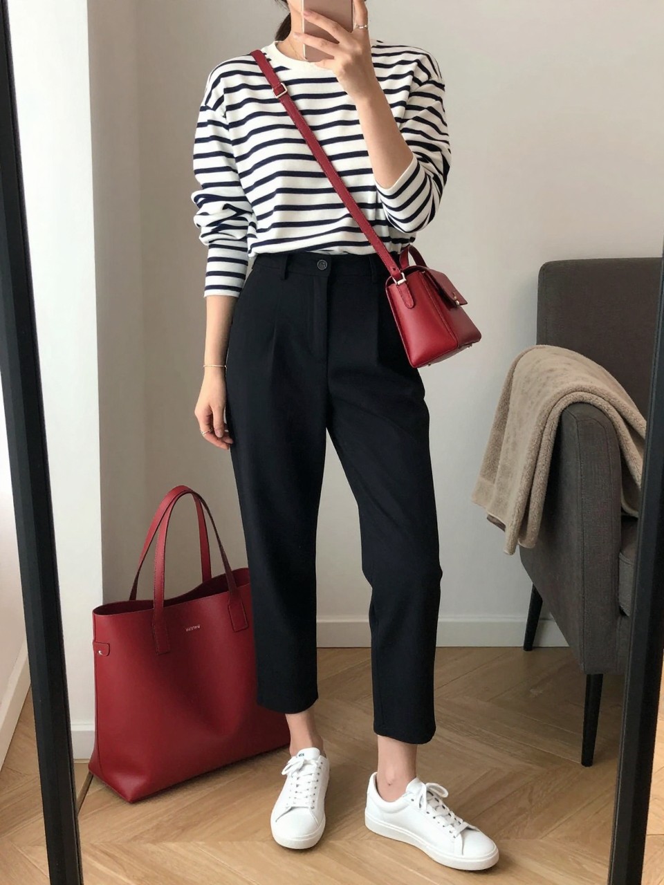 mirror selfie of a woman wearing the exact outfit described above, clearly showing a white and navy striped cotton Breton top, black cropped tailored trousers, white low-profile leather sneakers, a red leather crossbody bag, and hair in a sleek low ponytail, holding a phone in front of a mirror, cropped at NECK so the face does not show, with a relaxed casual selfie pose, in a stylish city apartment, soft indoor lighting, including tote bag resting on the floor and folded blanket on a chair, latest iPhone photo quality. (no watermarks on images)