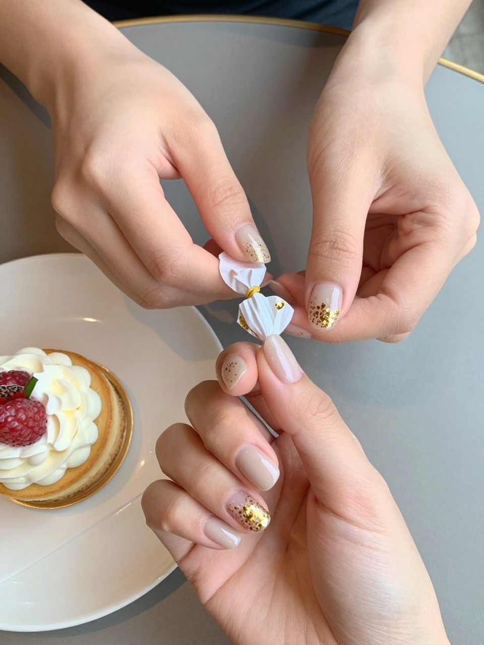 90-degree overhead top-down view of two feminine hands with oval medium nails.
The manicure features a soft beige base with gold confetti glitter concentrated along the tips.
Include glossy gel finish.

Hands are posed lightly holding a small wrapped candy.

Scene set on a café table with a small birthday dessert nearby.

Bright natural daylight with soft shadows, hyper-realistic lifestyle photography, sharp focus on nails, natural skin texture, realistic hands, no extra fingers.