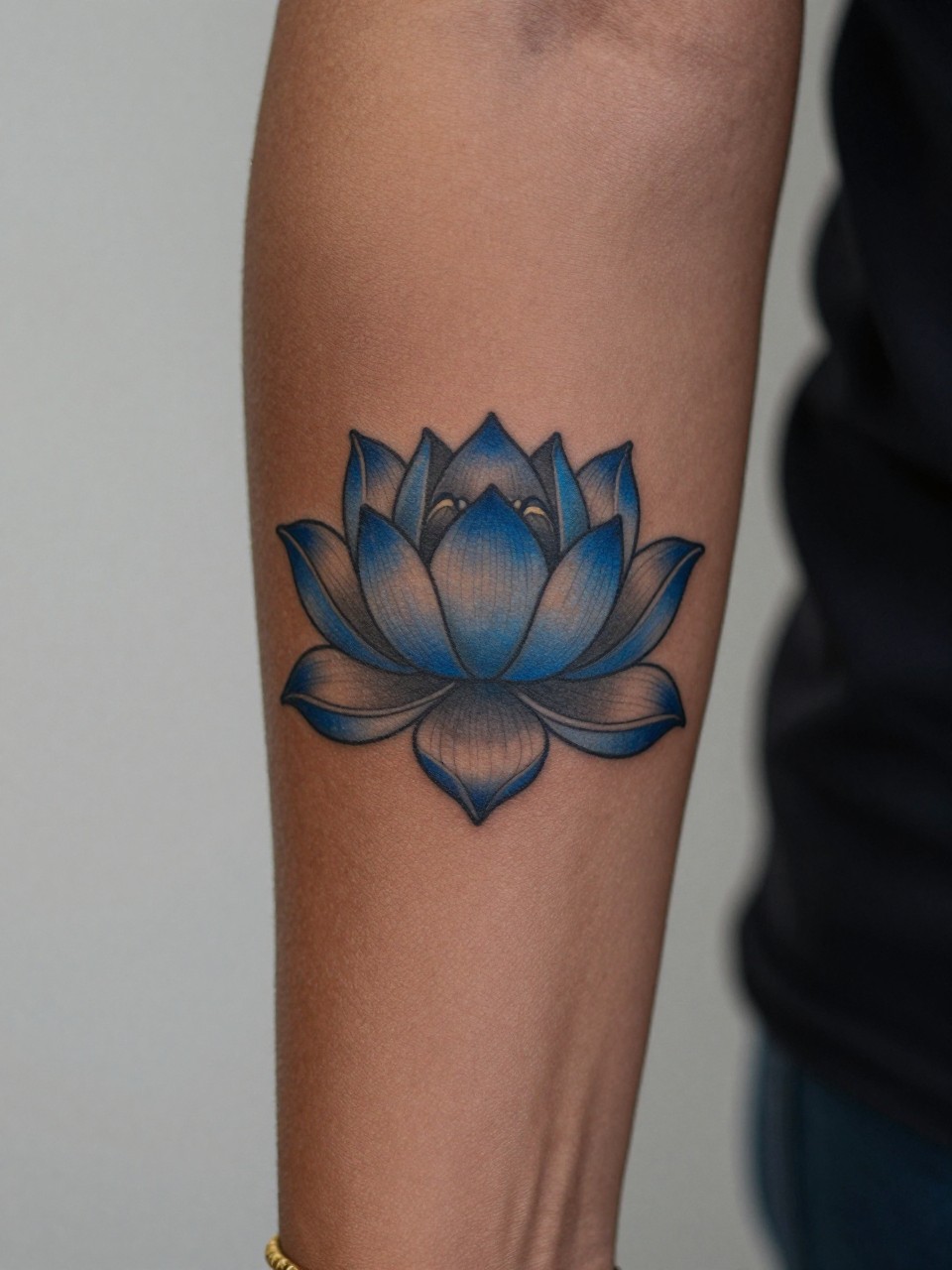 design-focused close-up of a blue lotus tattoo on deep brown skin at the forearm, rich blue petals, clean dark outline, soft shading, balanced floral design, realistic skin texture, gold bracelet visible, minimal blurred background, modern feminine tattoo inspiration photo (no watermarks on images)