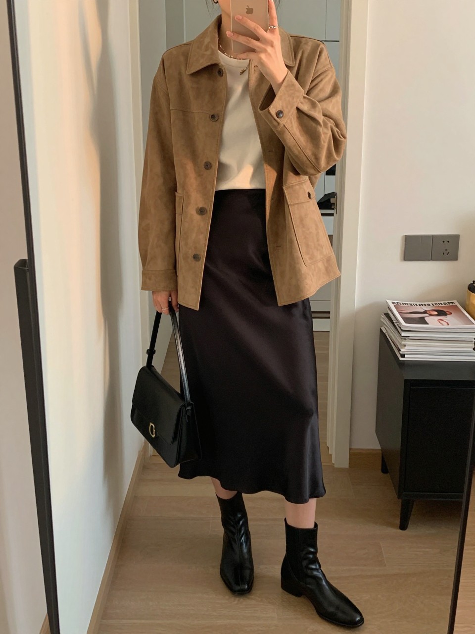 mirror selfie of a woman wearing the exact outfit described above, clearly showing a soft tan suede jacket, a black satin slip midi skirt, an ivory fitted tee, black flat ankle leather boots, and a structured black shoulder bag, holding a phone in front of a mirror, cropped at NECK so the face does not show, with a relaxed casual selfie pose, in a stylish city apartment, evening golden hour, including handbag hanging on a door handle and stack of fashion magazines on a side table, latest iPhone photo quality. (no watermarks on images)