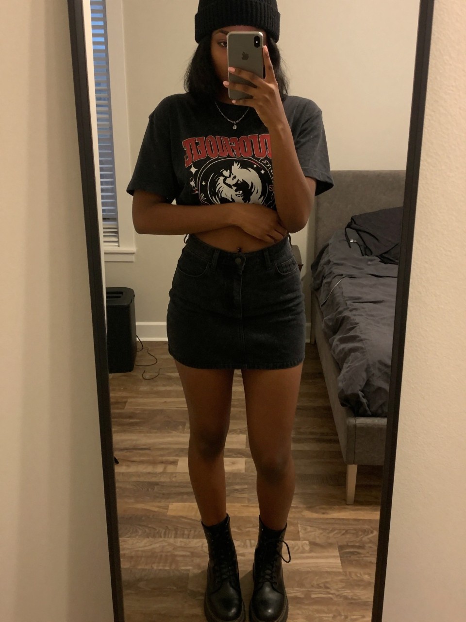 iPhone mirror selfie of a black woman in a graphic tee and black denim mini skirt with combat boots and a beanie, adjusting sleeve, cropped at NECK so face doesn't show, in a city apartment, warm moody lighting, iPhone photo quality.
