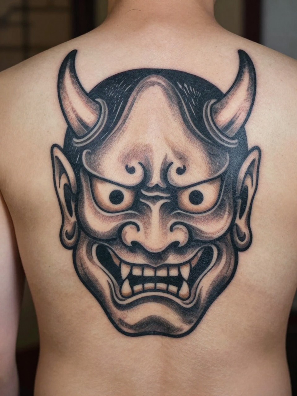 macro shot of a full back Japanese Oni mask tattoo on a man, bold demon face, curved horns, flame details, black and grey ink, thick traditional linework, dramatic shading, realistic skin texture, design-first back tattoo composition, soft neutral background (no watermarks on images)