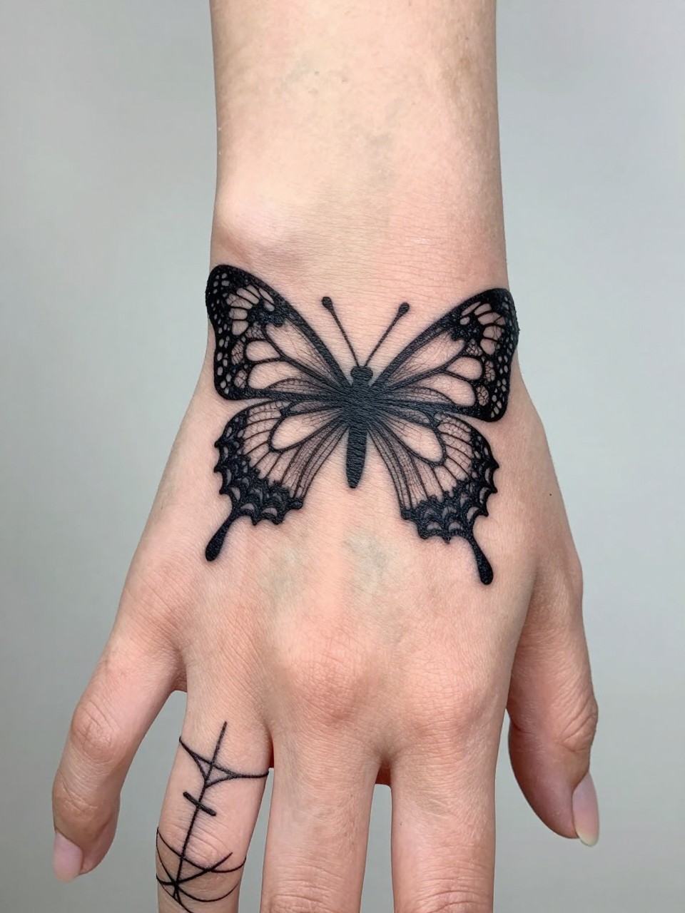 "Detailed shot of a wrist/hand tattoo: a butterfly tattoo with wings of the finest, most detailed black lace. The lace patterns connect to web designs on the fingers."