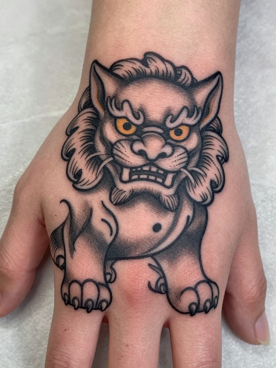 macro shot of a traditional Japanese Foo Dog tattoo on the back of the hand, guardian lion-dog face, curled fur details, black and grey ink, strong bold linework, smooth shading, realistic skin texture, design-first composition, soft neutral background, tattoo inspiration image (no watermarks on images)