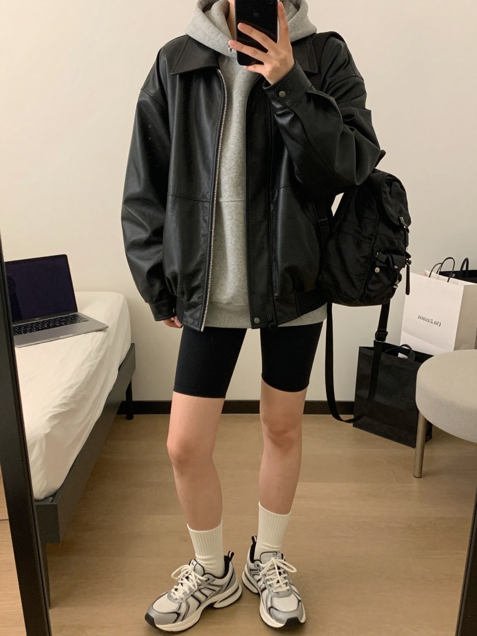 mirror selfie of a woman wearing the exact outfit described above, clearly showing an oversized black leather jacket with soft structure, heather gray fleece hoodie, black bike shorts, white crew socks with chunky sneakers, and a black nylon mini backpack, holding a phone in front of a mirror, cropped at NECK so the face does not show, with a relaxed casual selfie pose, in a minimal dressing room, warm moody lighting, including a laptop or tablet on the bed and shopping bags beside a chair, latest iPhone photo quality.