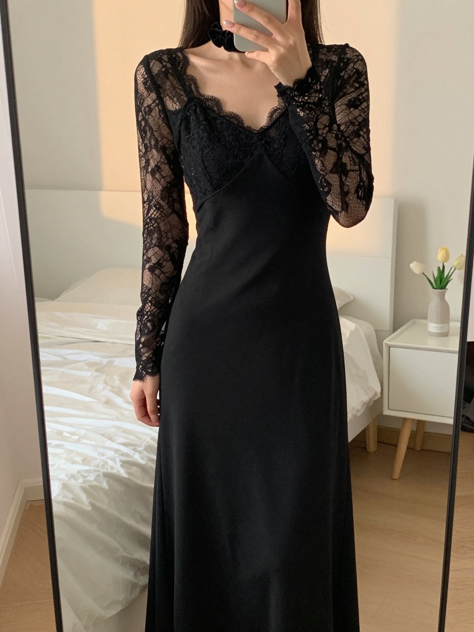 iPhone mirror selfie of a woman in a lace-sleeve black maxi, touching her velvet scrunchie, cropped at NECK so face doesn't show, in a feminine-themed bedroom, evening golden hour, iPhone photo quality.