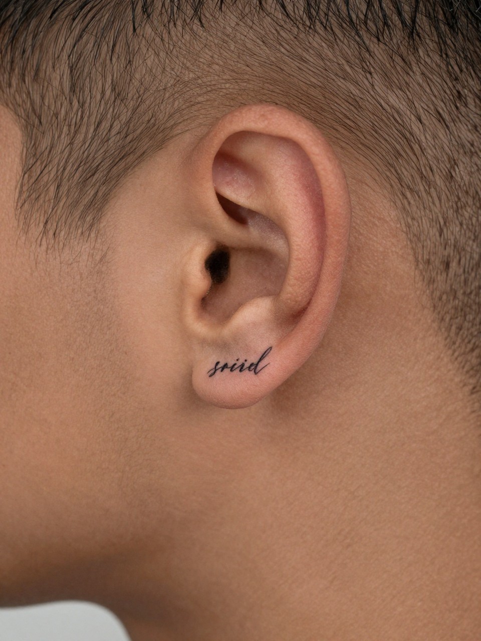 "macro shot of a script word tattoo behind the ear on rich brown skin, clean black ink, fine line cursive lettering, small elegant masculine design, realistic skin texture, sharp haircut edge, tight crop, design-first composition, minimal soft background, tattoo inspiration image (no watermarks on images)"