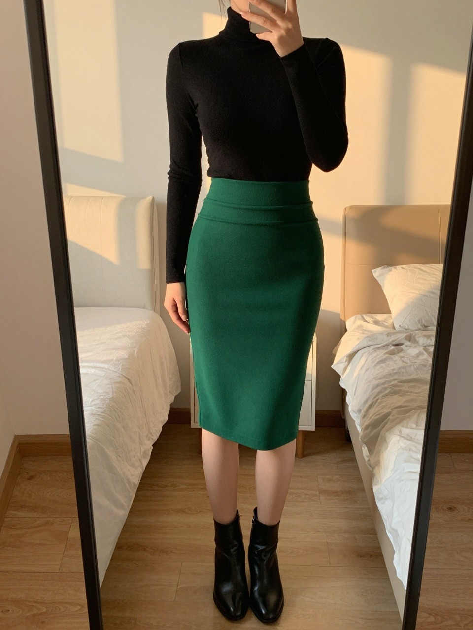 iPhone mirror selfie of a woman in a black turtleneck tucked into an emerald green pencil skirt with black ankle boots, standing with one hand in pocket, cropped at NECK so face doesn't show, in a feminine-themed bedroom, evening golden hour, iPhone photo quality.