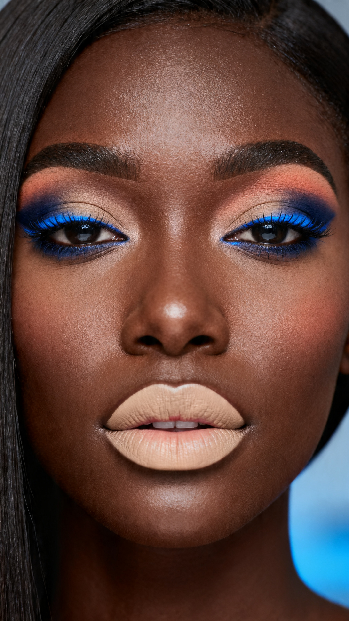 Super realistic close-up beauty portrait of a black woman wearing softly shaped neutral upper lids with a vivid cobalt and navy smoky lower lash line, top and bottom lash definition, muted peach-beige blush, satin nude beige lips, polished natural brows, realistic skin texture with a soft satin finish, shot from a front-facing close-up, with cool diffused beauty lighting that clearly reveals the bold lower-lash blue placement, lip texture, and smooth skin finish, sleek straight hair, blurred blue-gray background, feminine modern beauty photography, latest iPhone photo quality.