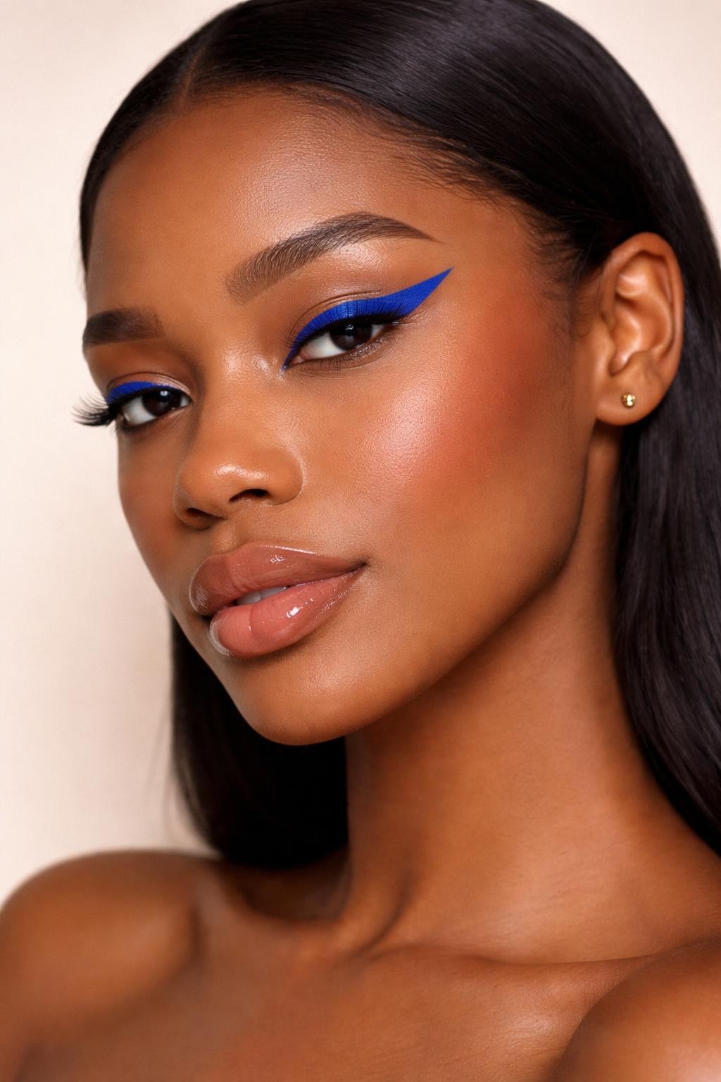 IMAGE_PROMPT: Super realistic close-up beauty portrait of a black woman wearing a bold vivid cobalt-blue winged liner on mostly clean neutral lids, soft peach-nude blush, beige-pink tinted lips, clean defined lashes, polished natural brows, realistic skin texture with a skin-like satin finish, shot from a three-quarter beauty angle, with diffused studio lighting that clearly reveals the crisp cobalt liner shape, warm cheek color, and natural lip finish, sleek hair tucked behind the ears, softly blurred cream background, feminine modern beauty photography, latest iPhone photo quality.