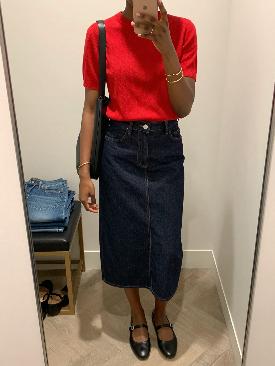 mirror selfie of a black woman wearing a red short-sleeve knit top with a dark denim midi skirt, black Mary Jane flats, a black leather shoulder bag, and slim gold bangles, clearly showing the same clothing items, colors, fabrics, and accessories listed in the Outfit Pieces section, holding a phone in front of a mirror, cropped at NECK so the face does not show, with a relaxed casual selfie pose, in a walk-in closet mirror, soft indoor lighting, including folded denim jeans on a bench and a pair of heels on the floor nearby, latest iPhone photo quality.