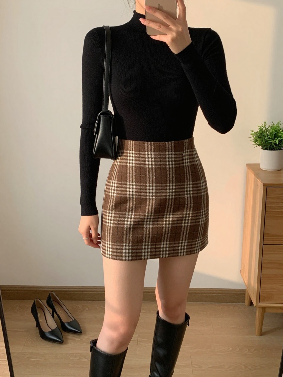 IMAGE_PROMPT: mirror selfie of a woman wearing a black fitted ribbed bodysuit, brown and cream plaid mini skirt, black leather ankle boots, black mini shoulder bag, and soft nude lipstick, holding a phone in front of a mirror, cropped at the NECK so the face does not show, relaxed casual selfie pose, in a minimal dressing room, evening golden hour, with heels on the floor nearby and small plant on a dresser. Latest iPhone photo quality.