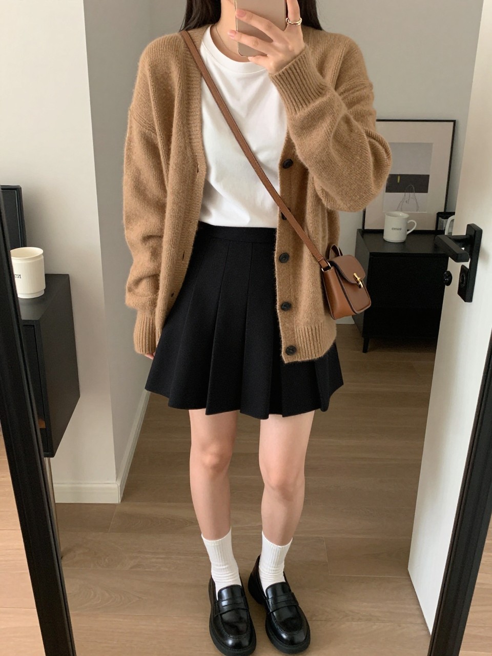 mirror selfie of a brown woman wearing a camel lightweight cardigan over a white fitted tee, black pleated mini skirt, black loafers, white ankle socks, and a small brown crossbody bag, clearly showing the same clothing items, colors, fabrics, and accessories listed in the Outfit Pieces section, holding a phone in front of a mirror, cropped at NECK so the face does not show, with a relaxed casual selfie pose, in a stylish city apartment, soft indoor lighting, including a coffee mug on a bedside table and a handbag hanging on a door handle, latest iPhone photo quality.