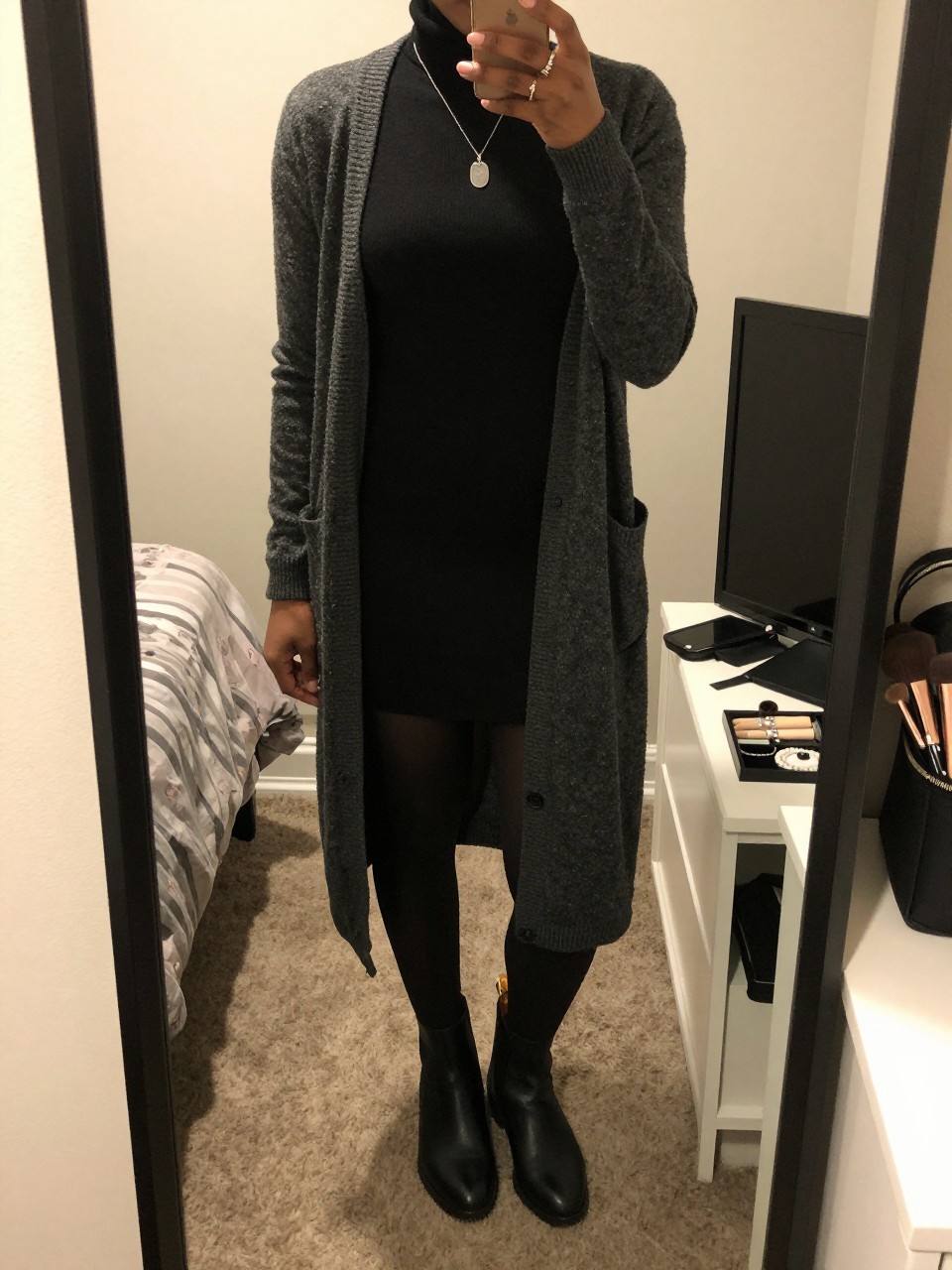 mirror selfie of a black woman wearing a charcoal long cardigan over a black turtleneck sweater dress, black opaque tights, black Chelsea boots, and a silver pendant necklace, clearly showing the same clothing items, colors, fabrics, and accessories listed in the Outfit Pieces section, holding a phone in front of a mirror, cropped at NECK so the face does not show, with a relaxed casual selfie pose, in a feminine-themed bedroom, warm moody lighting, including a makeup brush or cosmetic bag on the counter and a jewelry tray on the vanity, latest iPhone photo quality.