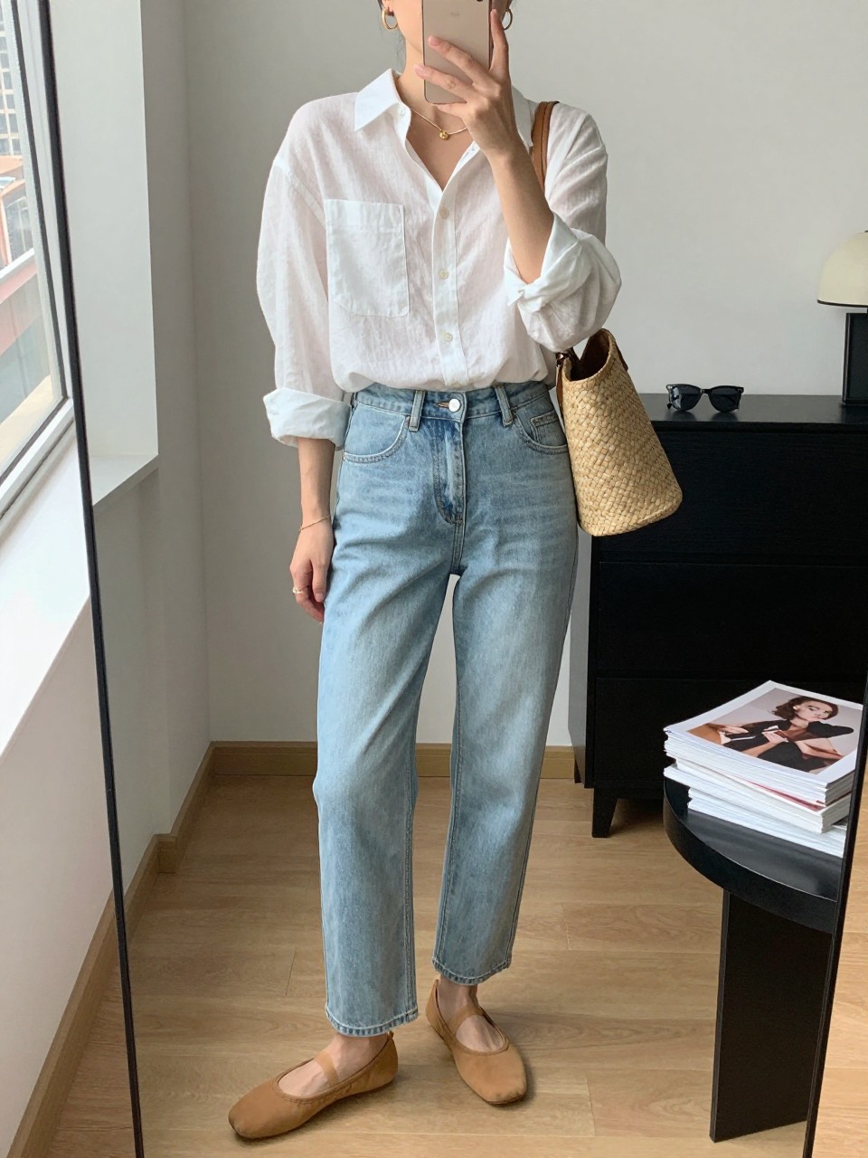 mirror selfie of a woman wearing a crisp white linen button-down shirt tucked into light wash straight-leg jeans, tan ballet flats, a small woven shoulder bag, and slim gold hoop earrings, clearly showing the same clothing items, colors, fabrics, and accessories listed in the Outfit Pieces section, holding a phone in front of a mirror, cropped at NECK so the face does not show, with a relaxed casual selfie pose, in a stylish city apartment, natural window light, including sunglasses placed on a dresser and a stack of fashion magazines on a side table, latest iPhone photo quality.