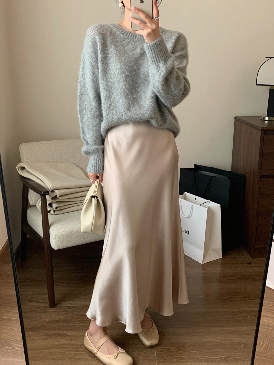 IMAGE_PROMPT: mirror selfie of a woman wearing a soft gray lightweight knit sweater, champagne satin maxi skirt, nude ballet flats, cream clutch bag, and pearl drop earrings, holding a phone in front of a mirror, cropped at the NECK so the face does not show, relaxed casual selfie pose, in a cozy bedroom mirror setup, warm moody lighting, with folded blanket on a chair and shopping bags beside a chair. Latest iPhone photo quality.