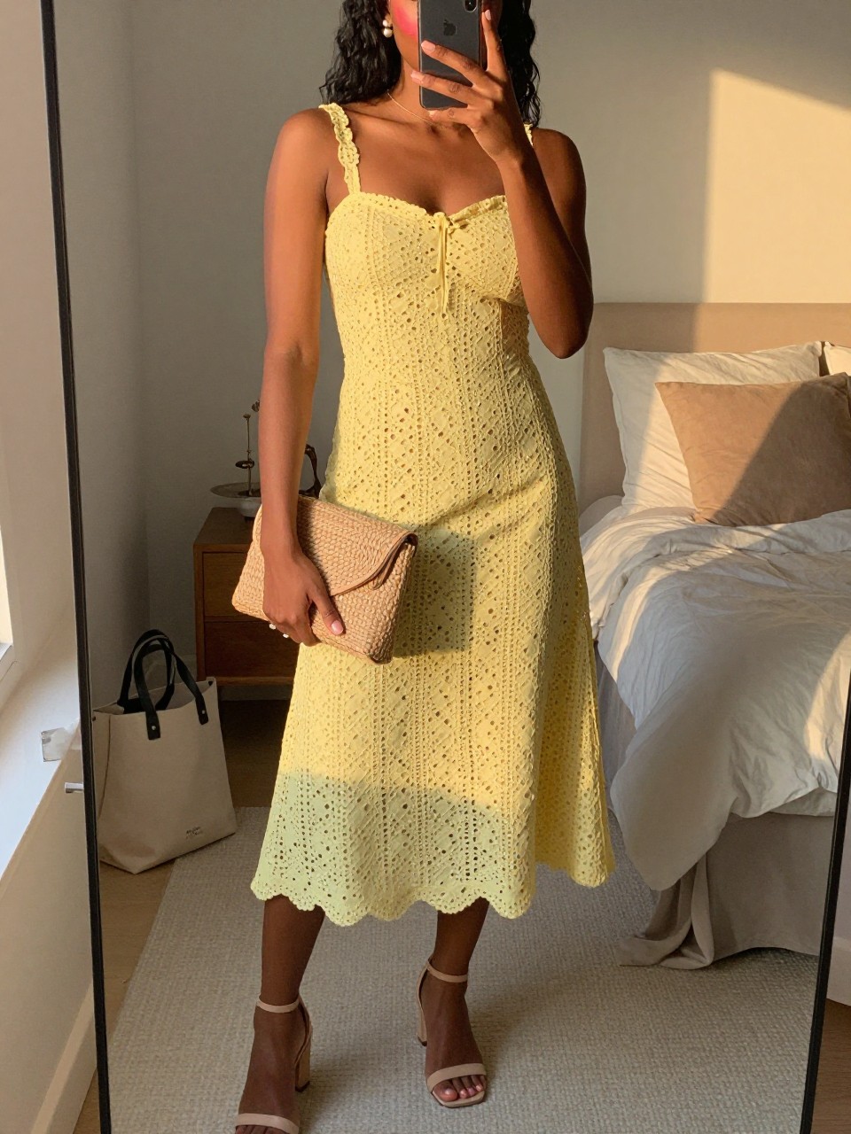 mirror selfie of a black woman wearing a butter yellow eyelet midi dress with nude block heel sandals, a natural woven clutch, pearl stud earrings, and rosy blush makeup, holding a phone in front of a mirror, cropped at NECK so face doesn't show, with a natural casual selfie pose, in a feminine-themed bedroom, evening golden hour, including small lived-in details like a tote bag on the floor and soft pillows on the bed, latest iPhone photo quality (no watermarks on images)