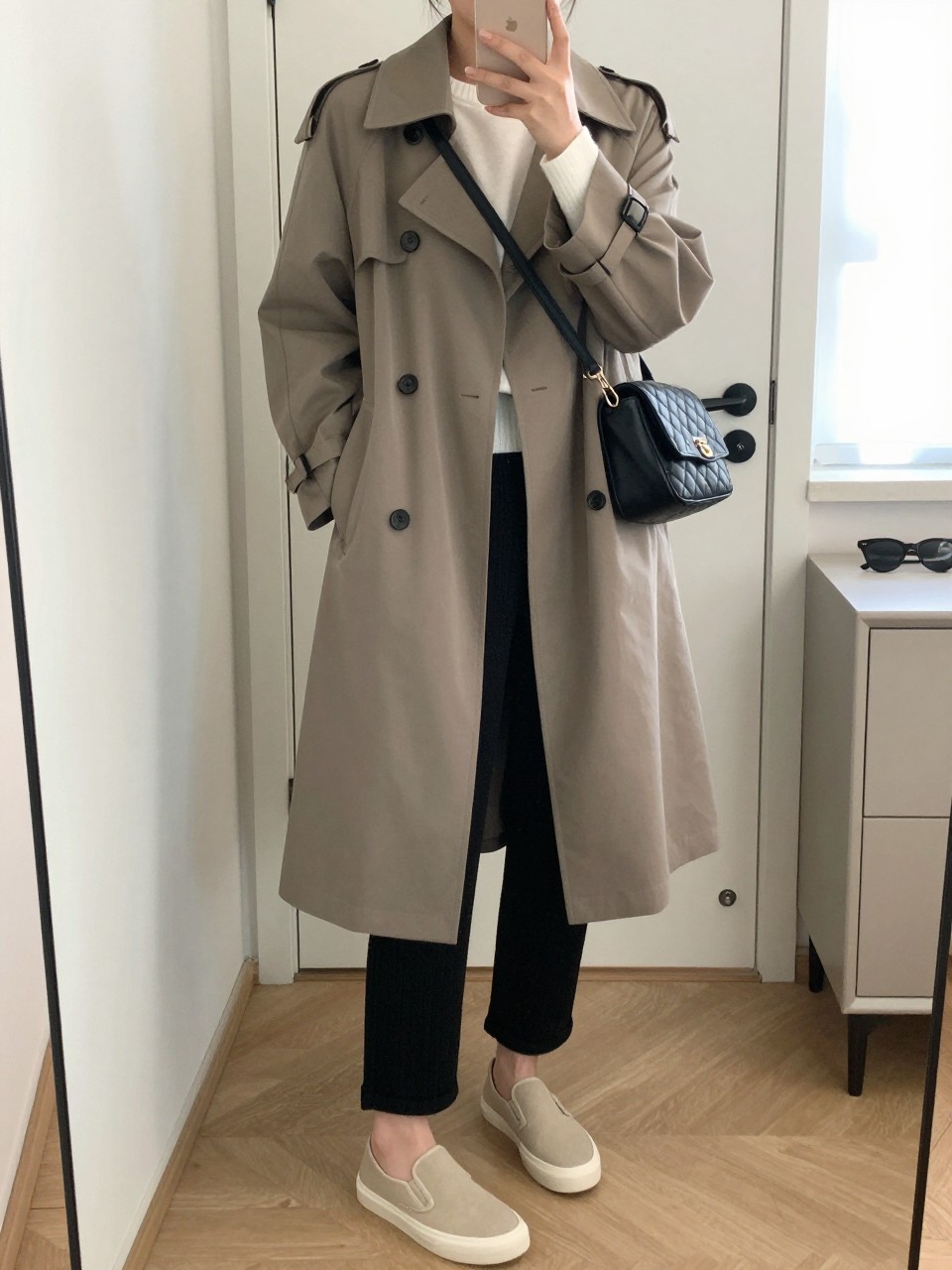mirror selfie of a woman wearing a taupe water-resistant trench coat over a white lightweight sweater, black knit straight-leg pants, beige slip-on sneakers, and a black quilted crossbody bag, clearly showing the same clothing items, colors, fabrics, and accessories listed in the Outfit Pieces section, holding a phone in front of a mirror, cropped at NECK so the face does not show, with a relaxed casual selfie pose, in a minimal dressing room, natural window light, including a handbag hanging on a door handle and sunglasses placed on a dresser, latest iPhone photo quality.